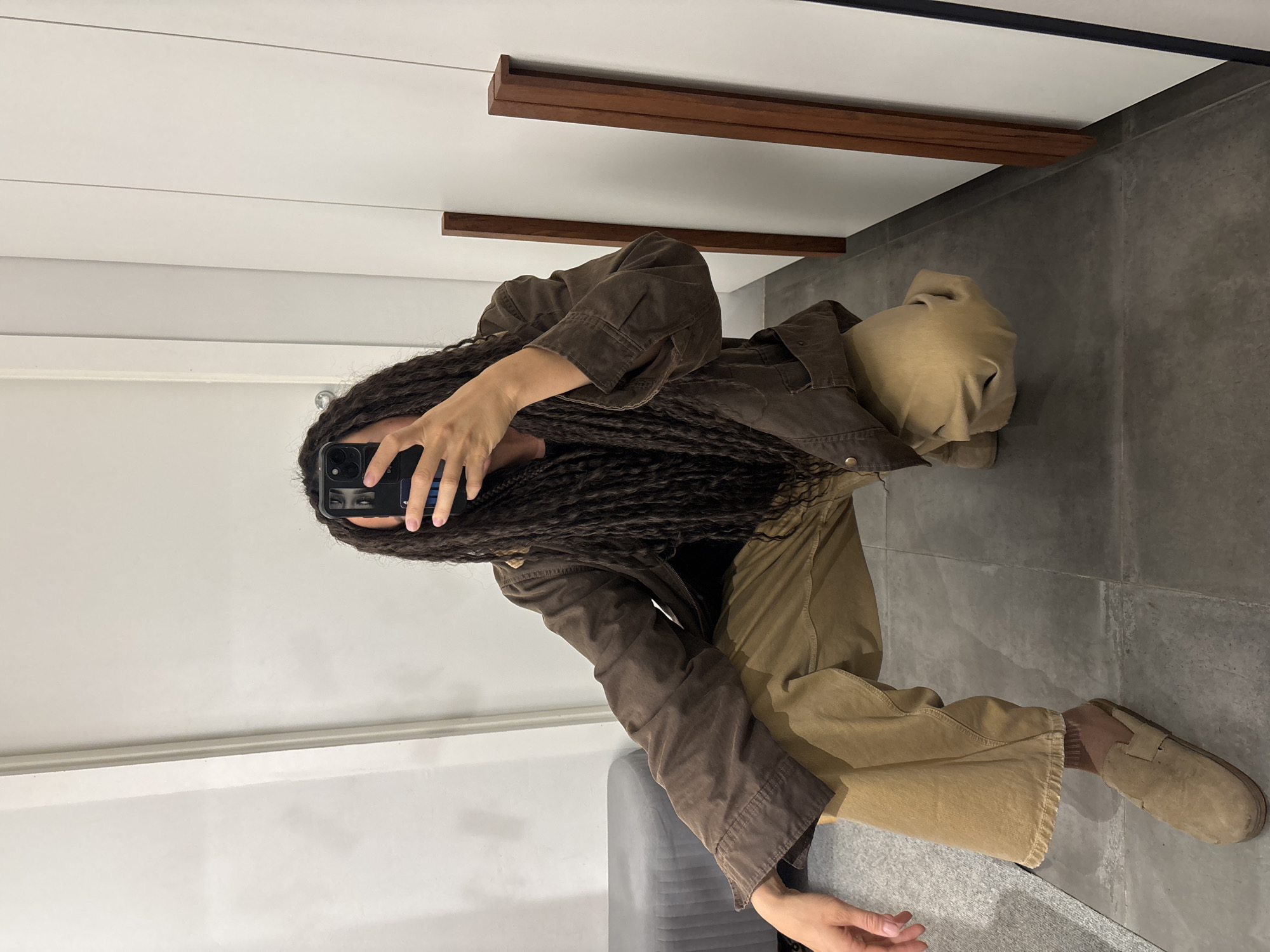 An indoor, medium shot captured in a mirror shows a person seated or kneeling, taking a selfie with a smartphone. The person's face is completely obscured by the black smartphone, which they hold up with both hands. Their long, dark braided hair cascades over their shoulders and arms. They are wearing a dark brown or olive-green jacket with visible seams and buttons, and loose-fitting, tan or khaki-colored pants. On their left foot, a light brown suede loafer or mule is visible, partially covering a ribbed sock.
The setting appears to be a modern interior, possibly a hallway, dressing room, or a contemporary living space. The floor is made of large, grey, concrete-like tiles, which also extend upwards to form a partial wall on the right. The background on the left features a white wall with horizontal paneling. Above this, a mirror reflects more of the white paneling and two parallel wooden beams or shelves mounted on the wall. A sliver of a grey, possibly upholstered, seat or table edge is visible in the lower center. The overall color palette is muted, with earthy tones of brown, tan, and grey, complemented by the clean white of the walls. No text is visible in the image.