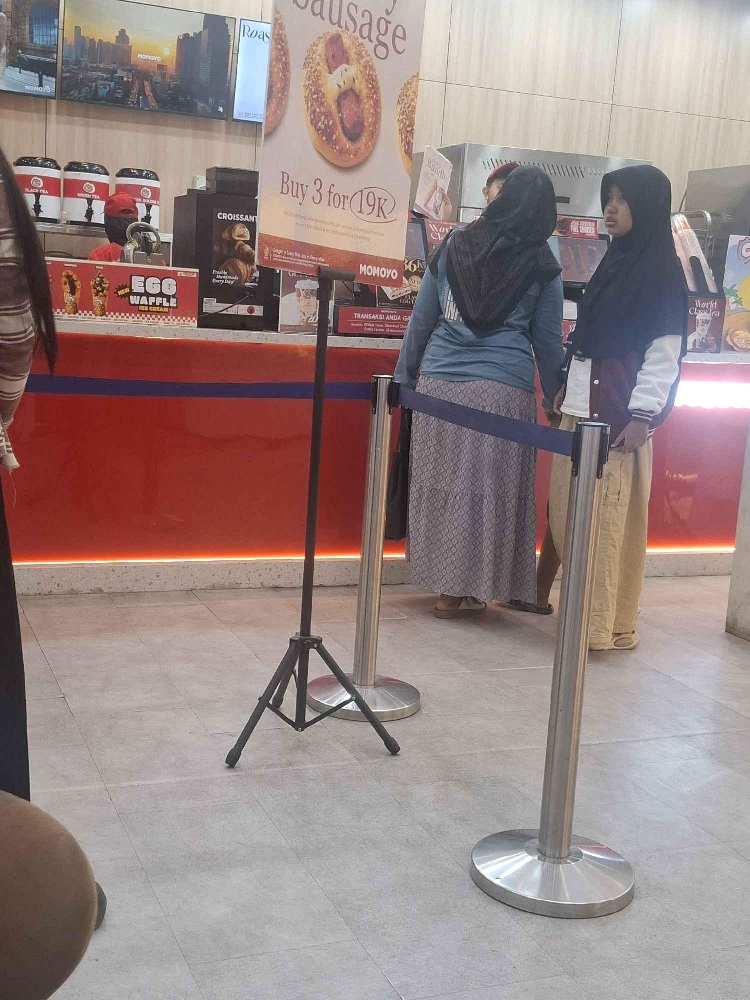 The image depicts an indoor scene, likely a cafe or food stall, with two women standing in line. One woman on the left wears a dark headscarf and a light blue long-sleeved shirt with a patterned skirt, and appears to be looking down. The other woman on the right wears a dark headscarf, a white and dark jacket, and light brown pants. Both women are facing towards a counter area. In the background, there is a large digital display with images of food and text, a red counter with various products displayed on shelves, and a queue barrier system. The flooring is tiled. The context of Cianjur, Indonesia is not visually evident in the image itself.