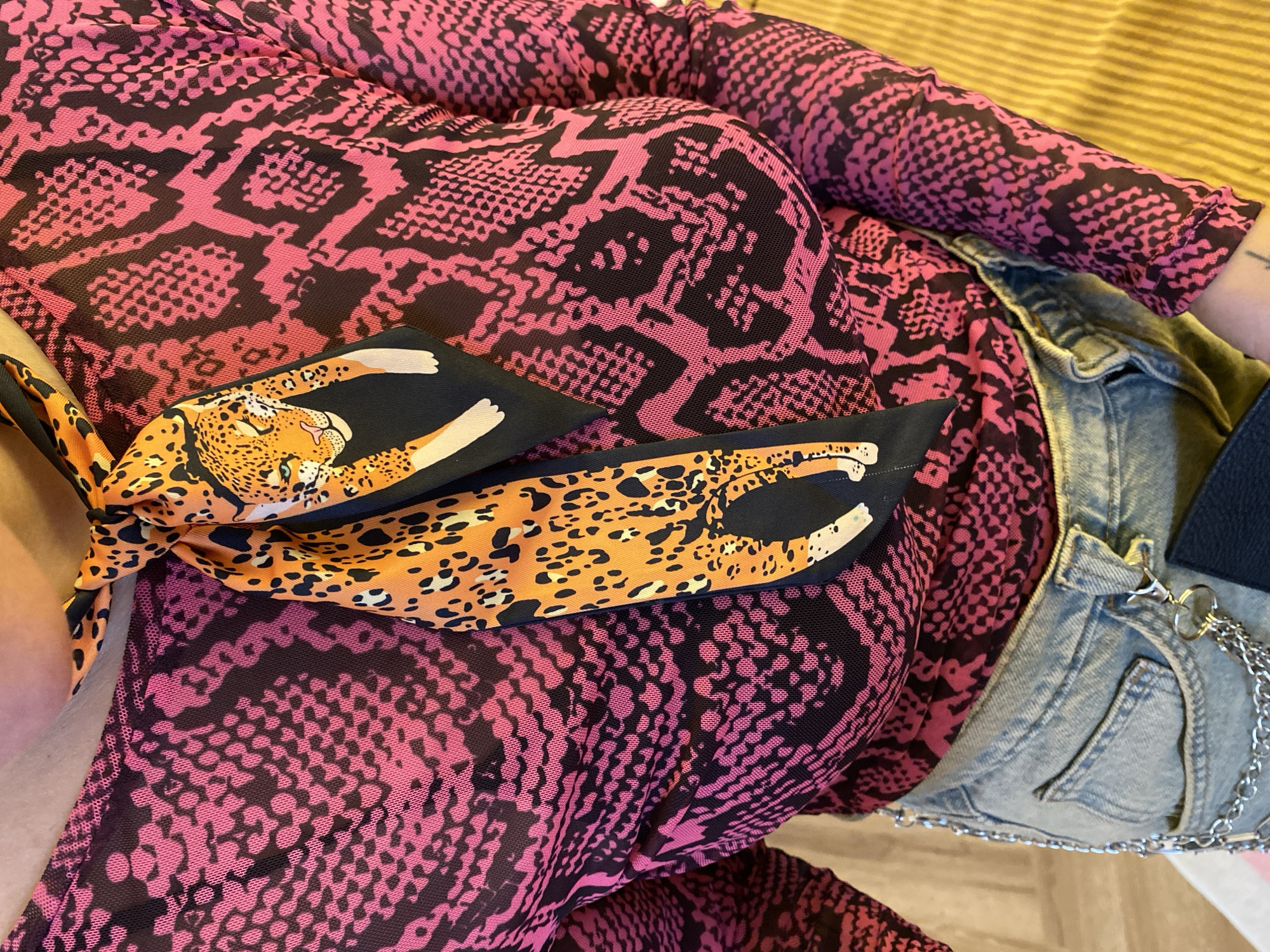 A close-up, medium-shot, high-angle, indoor photograph shows a person from the chest down, wearing a pink and black snake-print mesh top with a leopard-print scarf tied around their neck. The person is also wearing light-wash denim jeans with a silver chain attached to the belt loops. The background consists of a light-colored surface on the left and a textured yellow surface on the right. The lighting is bright and even. There is no indication of the location being Izhevsk, Russia.