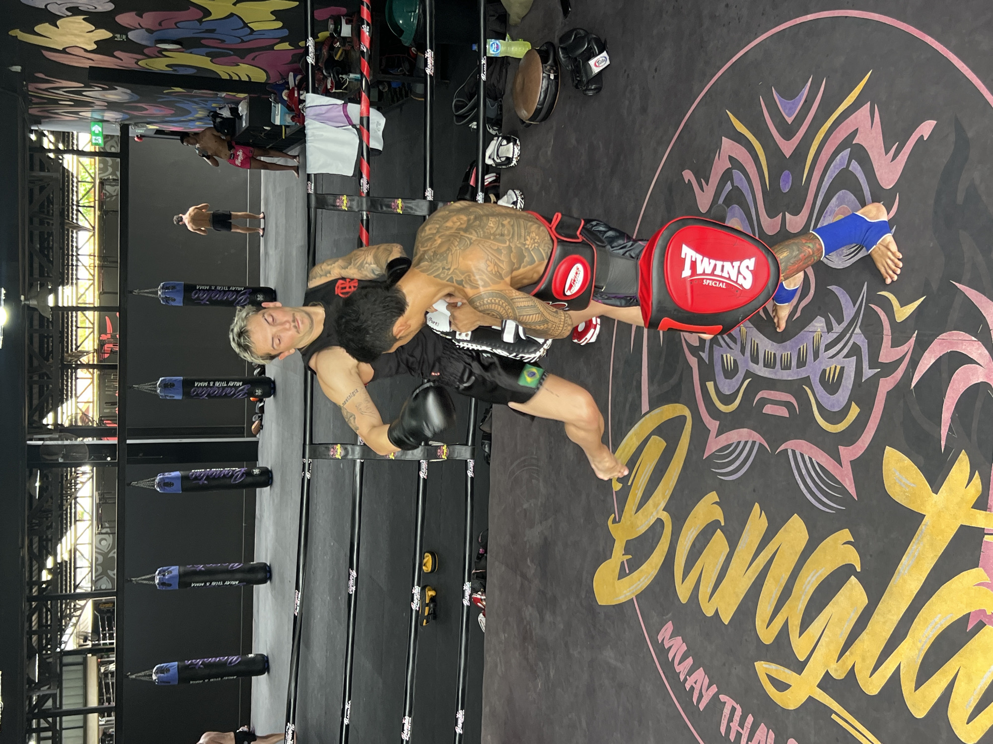 In a Muay Thai boxing ring in Choeng Thale, Thailand, two men are engaged in a training session. One man, wearing black shorts and a black tank top with a red logo, is throwing a punch with his left hand, which is covered by a black boxing glove. He has blond hair with dark roots and a mustache. The other man, wearing red and black Muay Thai shorts and blue ankle supports, is holding a red and black punch mitt to block the punch. He is heavily tattooed on his back, arms, and legs. The ring is marked with large yellow lettering that appears to read "Bangs" and a circular graphic with purple and pink abstract designs. In the background, several black and purple punching bags hang from the ceiling, and other individuals are visible in the gym. The overall environment is a brightly lit indoor training facility.