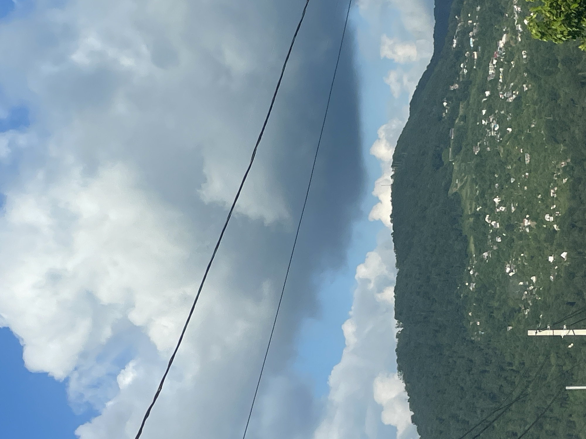 A wide shot captures a lush, green mountainside dotted with small white buildings, indicating a settlement on the slopes. The mountain is covered in dense trees, suggesting a natural, possibly forested, environment. The sky above is a mix of blue and dramatic, billowing clouds, with two dark, thin wires stretching horizontally across the upper portion of the frame. The scene appears to be a view of a residential area situated on a steep incline, likely in the mountainous outskirts of Sochi, Russia, given the presence of numerous houses on the green slopes. The overall impression is one of a scenic, somewhat rugged, landscape with human habitation integrated into the natural environment.