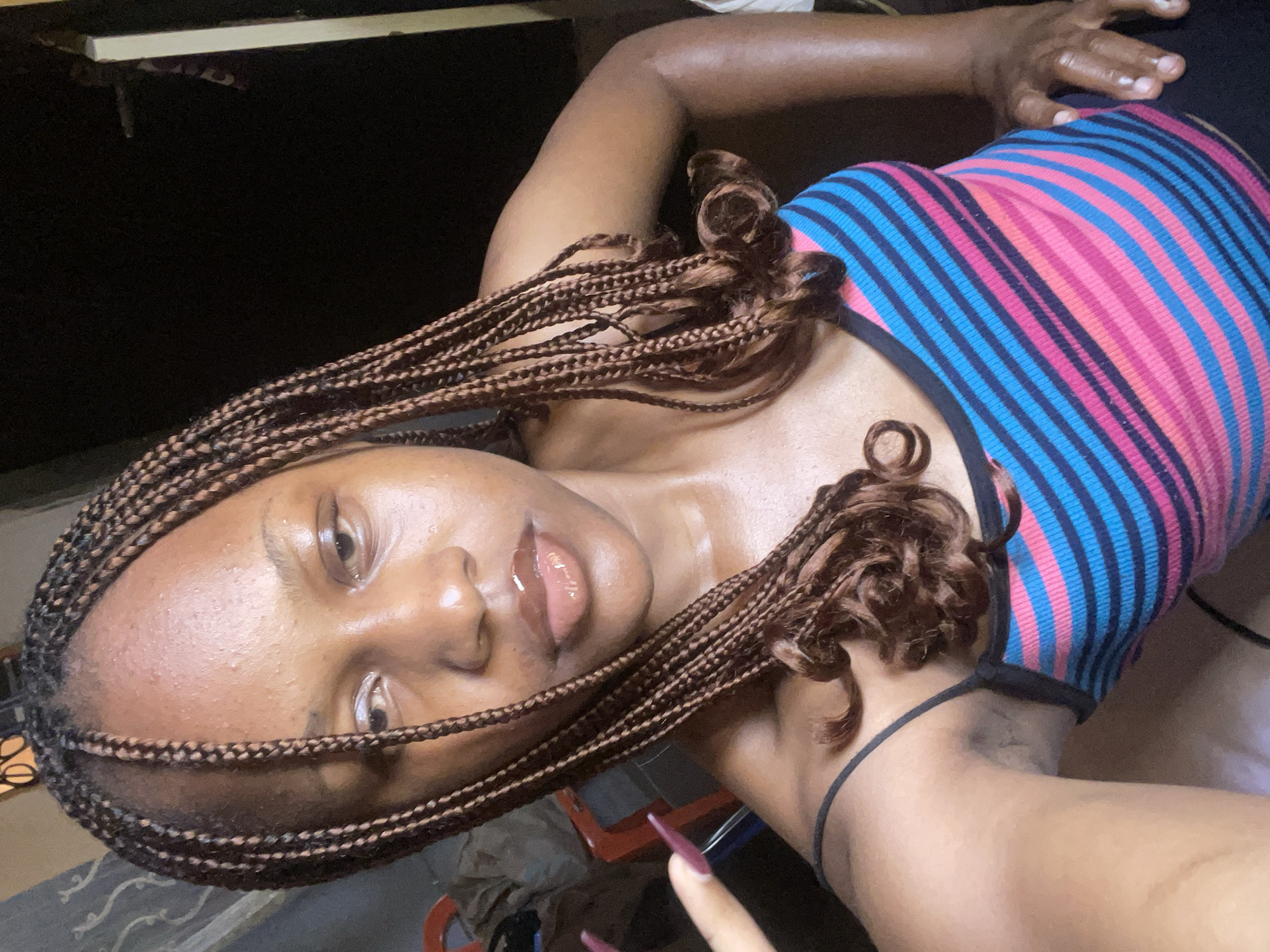 A young woman with braided hair and glossy lips is pictured from the chest up, looking towards the camera. She is wearing a striped tank top with pink, blue, and black bands. Her left hand, adorned with long, dark purple nails, is raised near her face. The background is dark and appears to be an indoor setting, possibly a room in Rafin Par, Nigeria, with hints of furniture and a doorway visible. The lighting suggests it is nighttime.