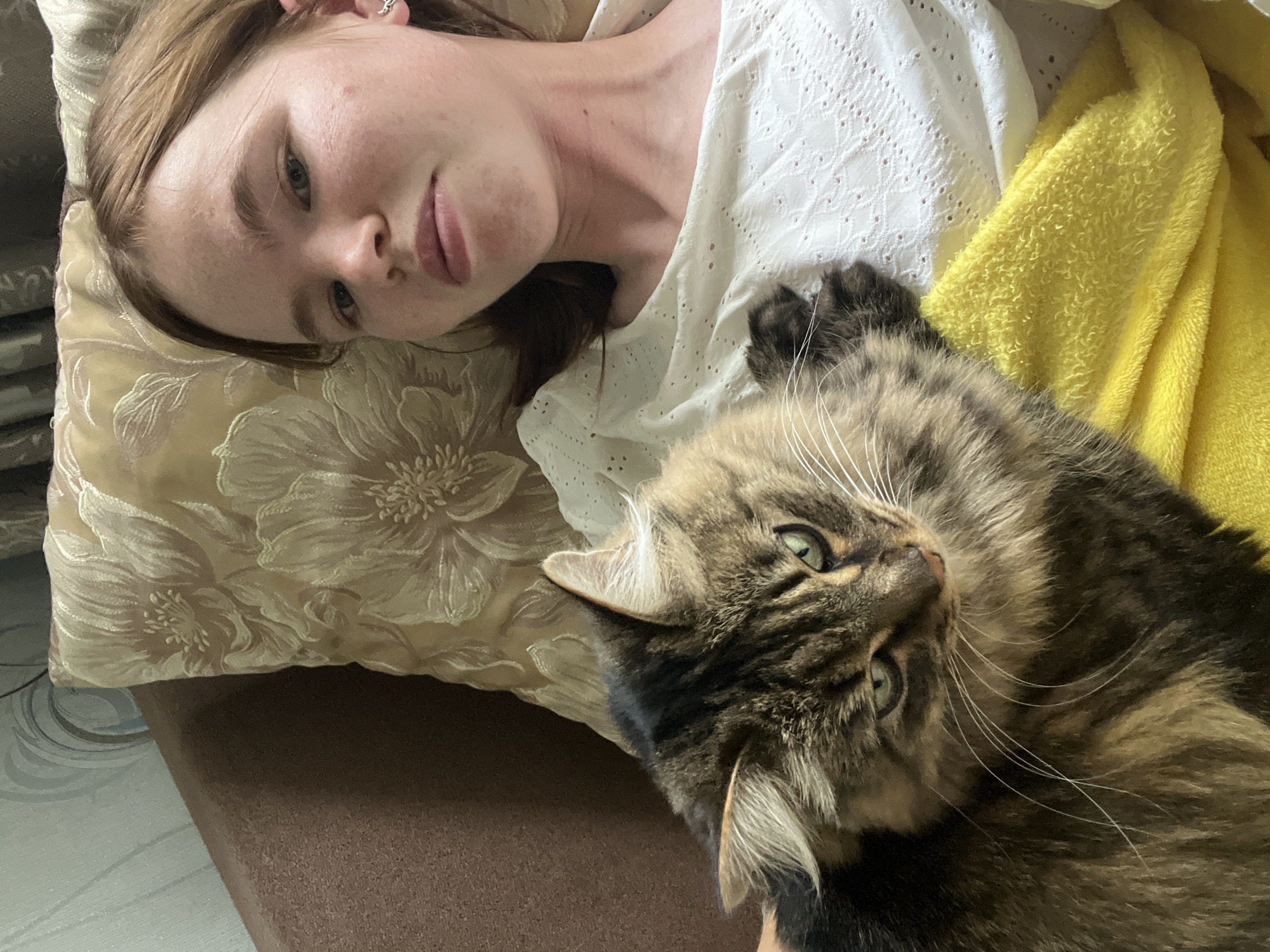 An overhead shot captures a young woman and a domestic cat resting together indoors in Chaikovsky, Russia. The woman, with light brown hair and fair skin, is lying on her back, her head resting on a cream-colored pillow with a raised floral pattern. She has light-colored eyes and a neutral expression, looking directly at the camera. She is wearing a white blouse with a delicate eyelet pattern. A small, silver-colored earring is visible in her left ear.
Beside her, a long-haired tabby cat with brown, black, and lighter fur is also resting, its head tilted and its green eyes looking towards the camera. The cat has prominent white whiskers. A bright yellow, textured blanket or towel is draped near the cat and the woman's shoulder. Both subjects appear relaxed. The background includes a dark brown textured surface, likely a couch or bed, and a small portion of a light-colored floor with a subtle pattern is visible in the lower left corner. The lighting suggests it is daytime. No text is visible in the image.
