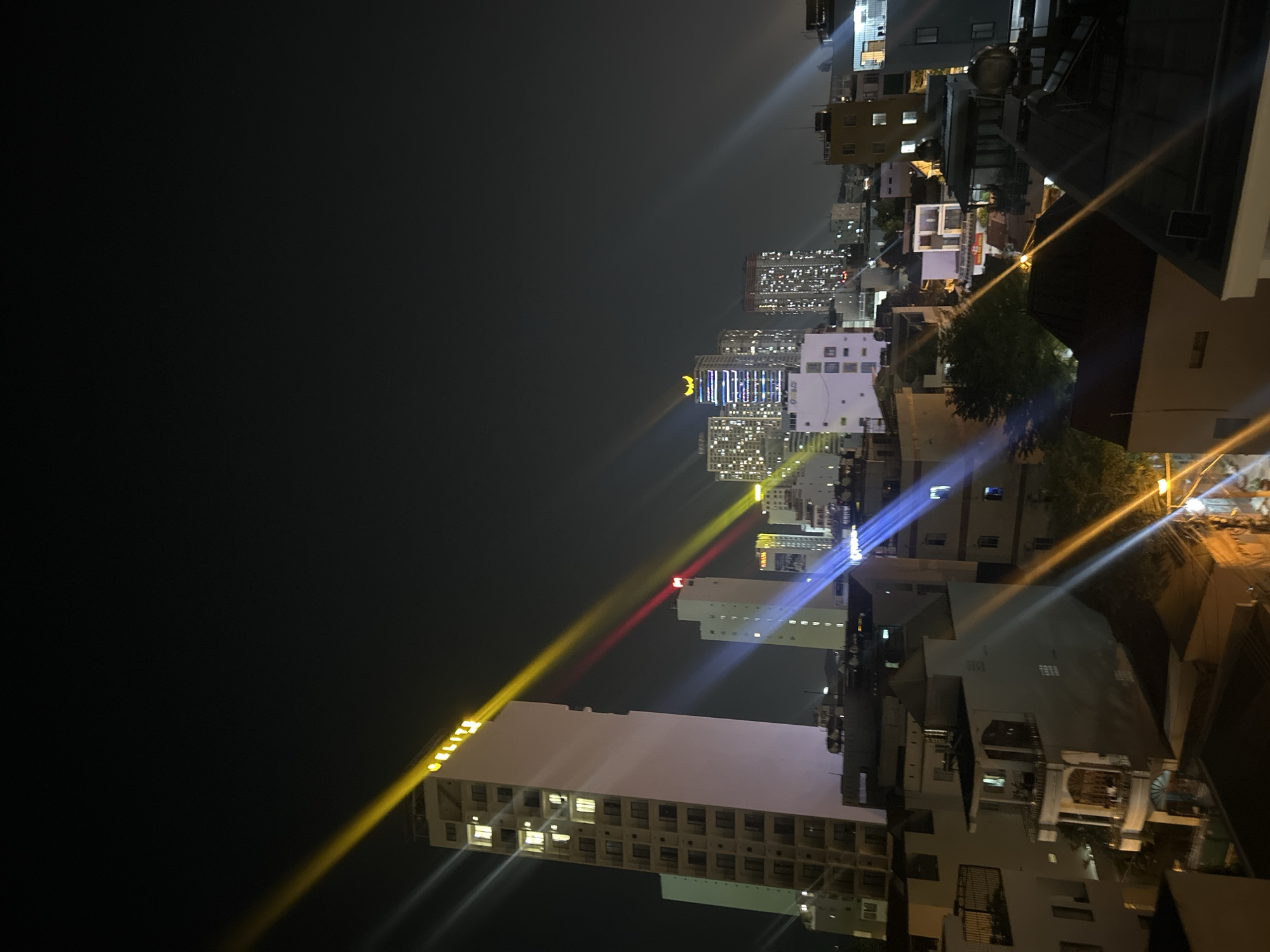 The image shows a night view of Bắc Nha Trang, Vietnam. High-rise buildings dominate the skyline. Lights from windows and streetlights illuminate the scene, creating lens flares. The sky is dark and clear. The photo seems to be taken from a high vantage point, likely a rooftop or high floor of a building, offering a panoramic view of the city.