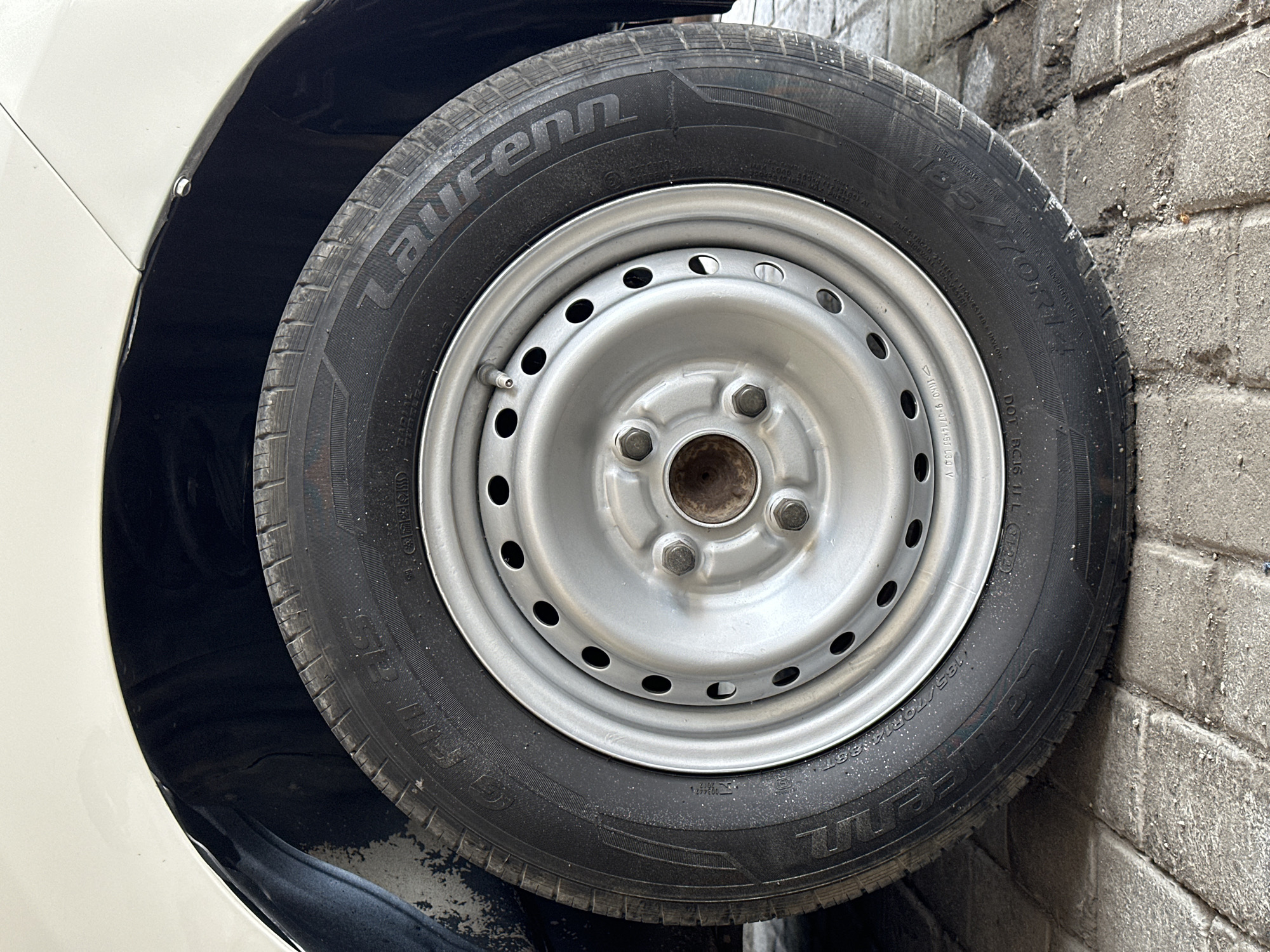 The image shows a close-up, angled shot of a car tire and wheel, positioned in what appears to be an outdoor setting with a paved ground surface made of bricks. The car is white, with a portion of its fender visible above the tire. The tire itself is black and has white lettering, indicating the brand "Laurenn" and the size "185/70R14". The wheel is a silver-colored steel rim with four visible lug nuts securing it to the hub. The tire shows signs of use, with some dirt and scuff marks. The background is dark, likely the wheel well of the car, and the ground is a light-colored brick pattern. There are no people or animals visible in the image, and no specific landmarks or indicators point to Bulakrejo, Indonesia.