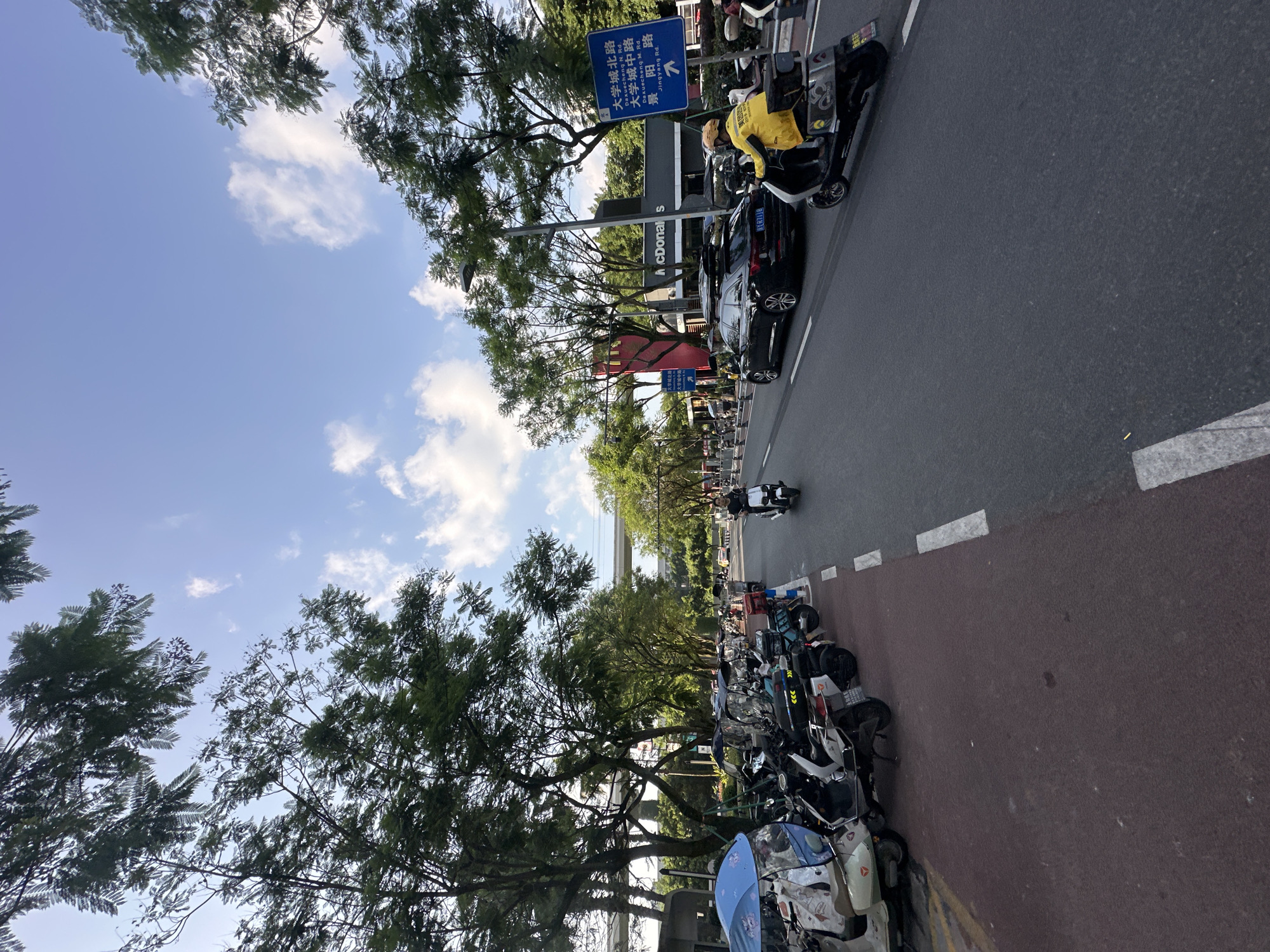 The image depicts a street scene in the city of Chongqing High-tech Zone, China. The time of day appears to be late afternoon, with a bright sky and scattered clouds. The street is paved with asphalt and has white lane markings.
On the left side of the street, a row of parked electric scooters and motorcycles are visible, some covered with tarps and one with a blue umbrella. A red pedestrian walkway runs alongside the parked vehicles.
In the road, several vehicles are present. A black sedan with a roof rack is driving in the right lane. A person on a scooter, wearing a bright yellow jacket and helmet, is also in the right lane, moving towards the viewer. Another person on a white scooter is seen further ahead in the same lane, also moving towards the viewer.
On the right side of the road, a blue sign with Chinese characters and an arrow indicates directions. A McDonald's restaurant is visible in the background. Tall green trees line both sides of the street, providing shade and creating a canopy effect. An elevated structure, possibly an overpass or monorail, is visible in the distance, stretching across the scene. The overall environment suggests a bustling urban area with a mix of transportation modes and commercial establishments.