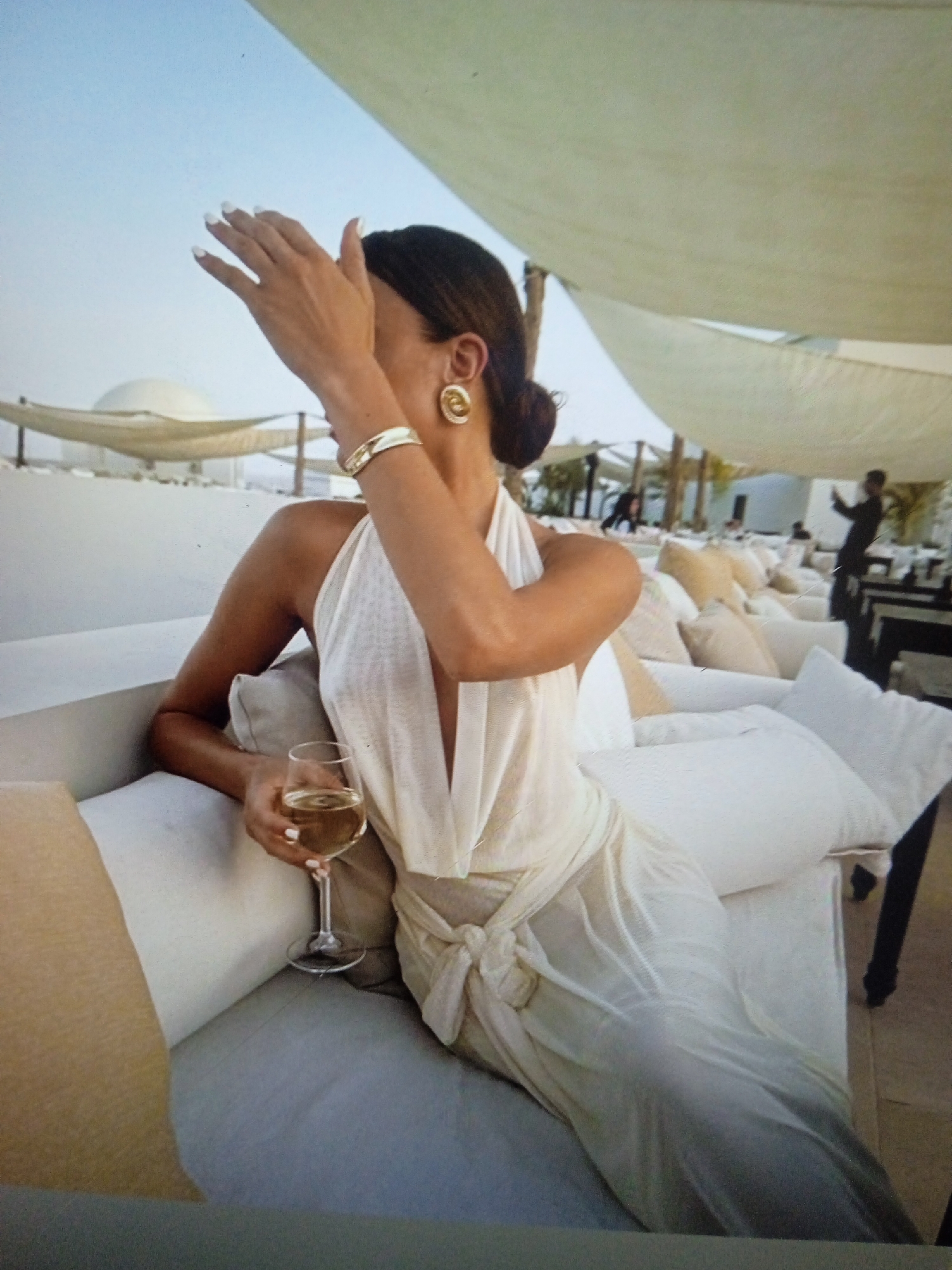 The image shows a woman in a white dress with a halter top, seated on a white sofa. She is holding a glass of white wine in her right hand and has her left hand raised to her face. She is wearing a bracelet and large hoop earrings. The background appears to be an outdoor seating area with more white sofas and cushions, covered by a light-colored canopy. In the distance, there are people and tables. The setting is a bright, sunny day with a clear sky. The image does not depict any location in Chernivtsi, Ukraine.
