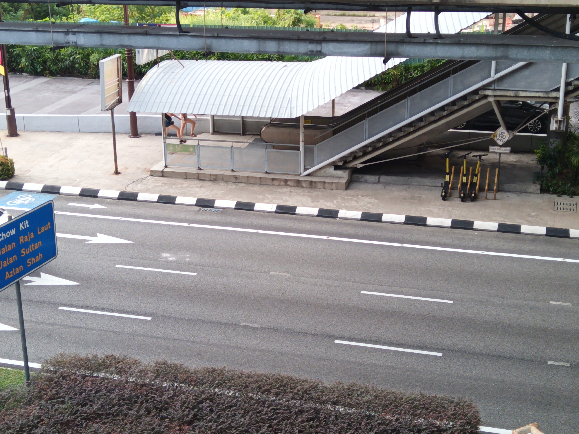 The image shows an outdoor scene in Kuala Lumpur, Malaysia, likely near a transportation hub. In the foreground, a road is visible with white lane markings and a blue directional sign indicating "Chow Kit", "Jalan Raja Laut", "Jalan Sultan", and "Azlan Shah". Adjacent to the road is a strip of dark green and purple foliage.
Above the road, there is a pedestrian walkway with a black and white checkered curb. A covered walkway, possibly an entrance to an escalator or stairs, is visible. Two individuals, one appearing to be wearing shorts and a t-shirt, are standing under this covered area. An escalator and a staircase are part of the infrastructure, leading upwards.
Further back, a metal structure, likely an overhead walkway or part of a larger building, is prominent. The background reveals lush green trees and vegetation. Several yellow and black electric scooters are parked in a designated area beneath the escalator structure. A car is partially visible in the background, parked under the elevated structure. The overall scene suggests a public area designed for pedestrian and vehicular traffic, possibly near a transit station or a busy street in the city.