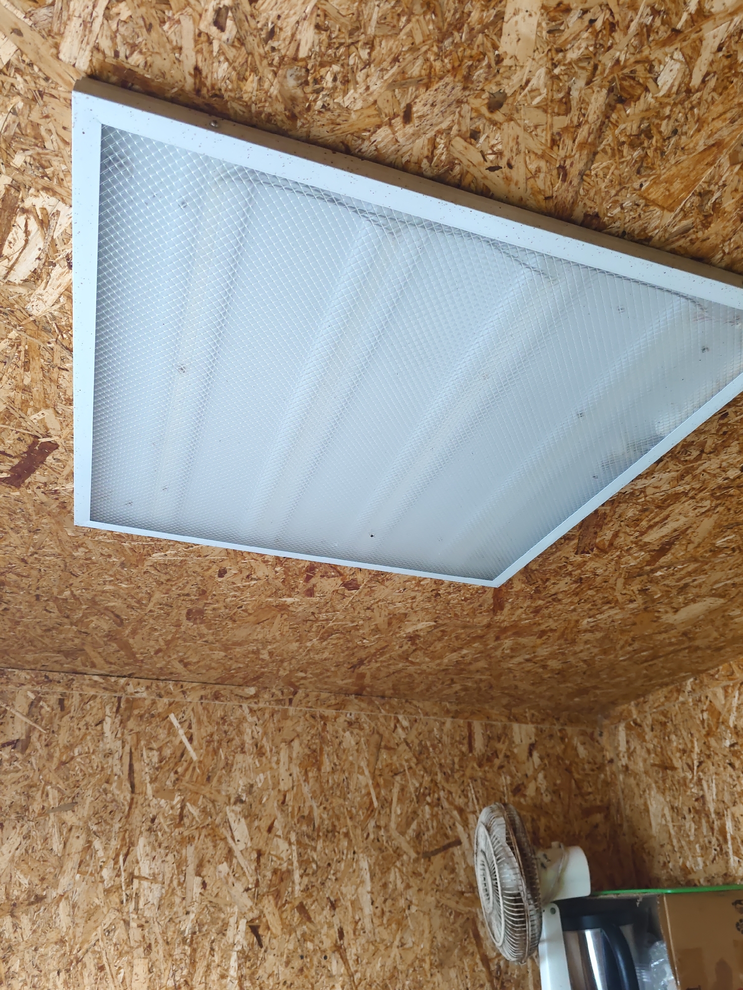 A fluorescent light fixture is mounted on a ceiling made of oriented strand board (OSB). The light fixture is rectangular with a white frame and a translucent, textured diffuser. Below the light fixture, on a wall also made of OSB, a white electric fan is visible, positioned on a shelf. The overall environment appears to be utilitarian, possibly a workshop or storage area.