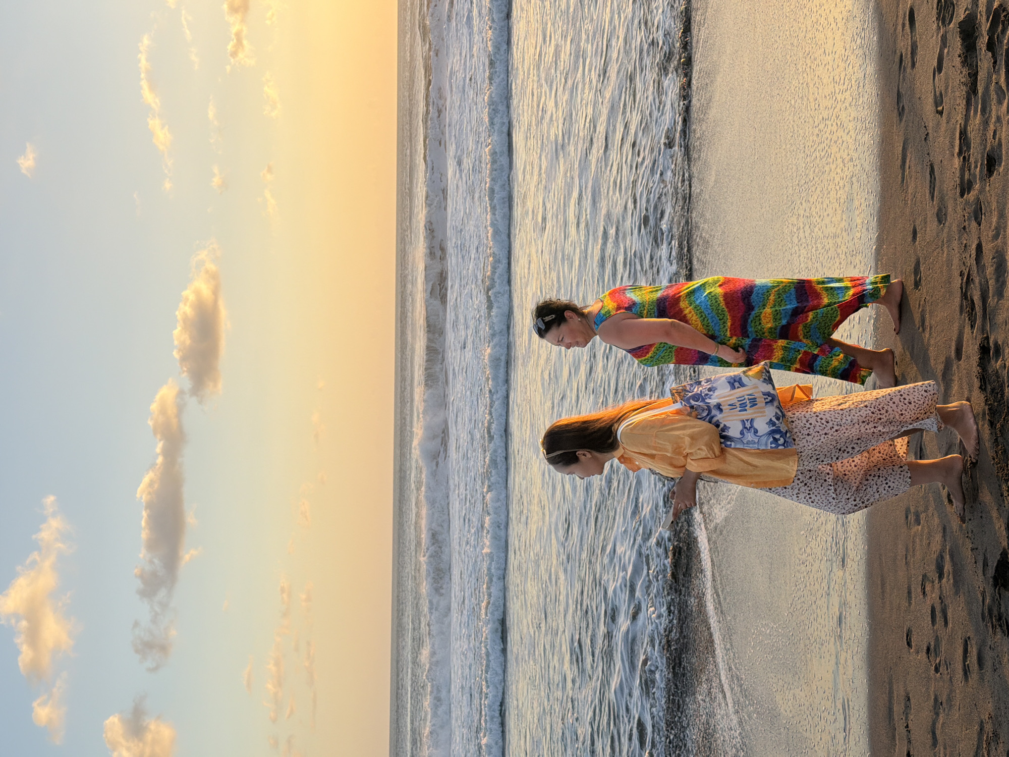 In North Kuta, Indonesia, two women are captured in a full-length shot walking barefoot on a sandy beach during golden hour. The woman on the left, facing left, wears an orange long-sleeved shirt and patterned pants, carrying a blue and white tote bag over her shoulder and holding a smartphone. The woman on the right, also facing left, is dressed in a vibrant, multi-colored tie-dye maxi dress. They are positioned on the dark sand of the beach, with the ocean and waves to their left. The ocean water is a deep blue, and the waves are breaking. The sky above transitions from a pale blue at the top to a warm orange and yellow hue near the horizon, indicative of a sunset, with scattered white and yellow clouds. The sun is setting behind the ocean, casting a golden light across the scene. The foreground shows the sandy beach with footprints.