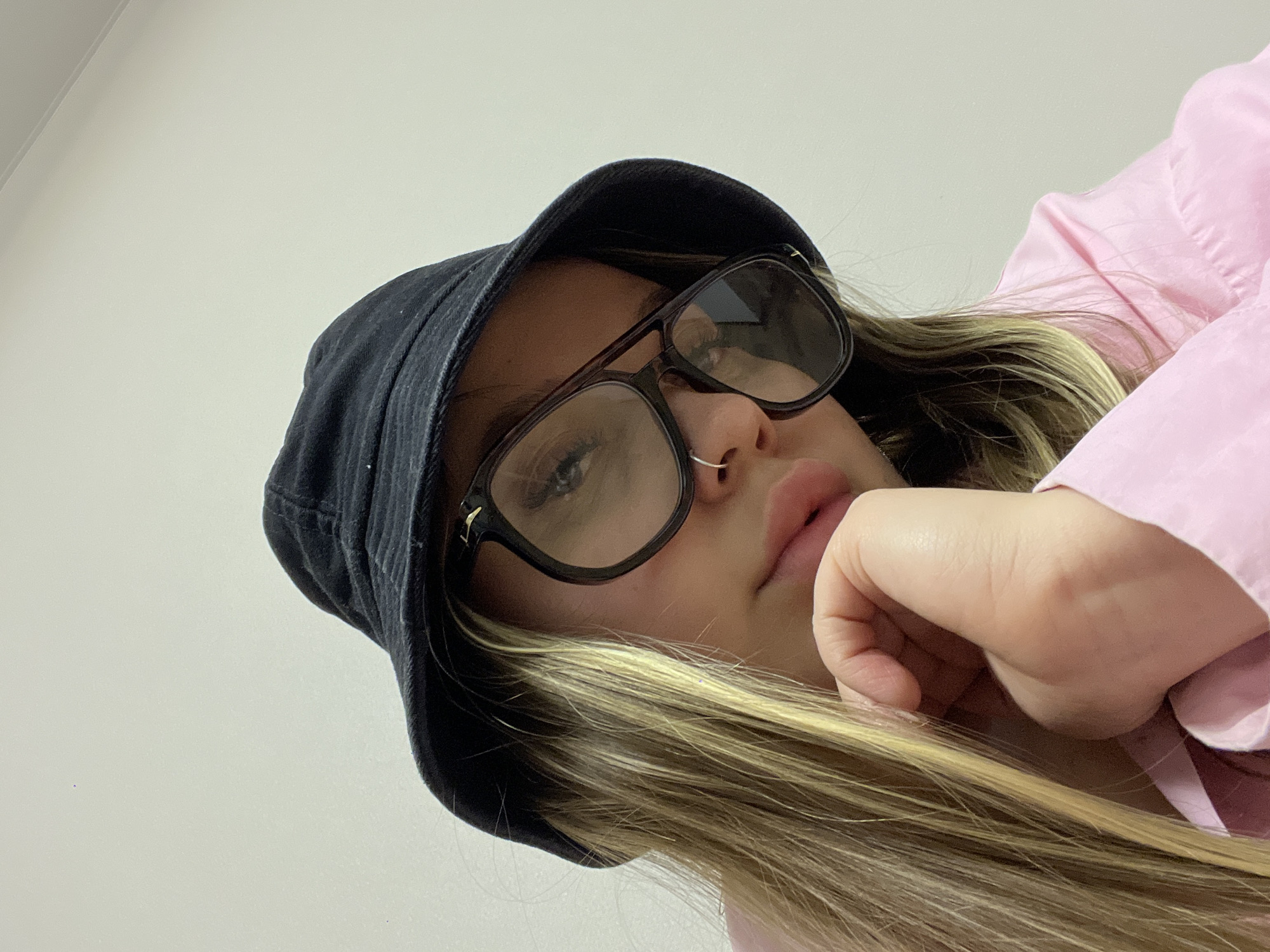 A young individual, likely female, is captured from a low-angle perspective, looking upwards towards the camera. The subject wears a black bucket hat and large, dark-rimmed glasses, which obscure most of their eyes. Reflections are visible on the glass lenses. A small silver hoop nose ring adorns their right nostril. Long, wavy hair with blonde highlights and darker roots falls from beneath the hat. One hand, with fingers slightly curled, rests near their mouth and chin area. A light pink, collared shirt is visible on the right side of the frame. The background consists of a plain, light-colored wall, with a subtle line indicating a corner or ceiling joint in the top left. The lighting is soft and even, suggesting an indoor setting. No text is visible in the image. The person's expression appears neutral or contemplative.