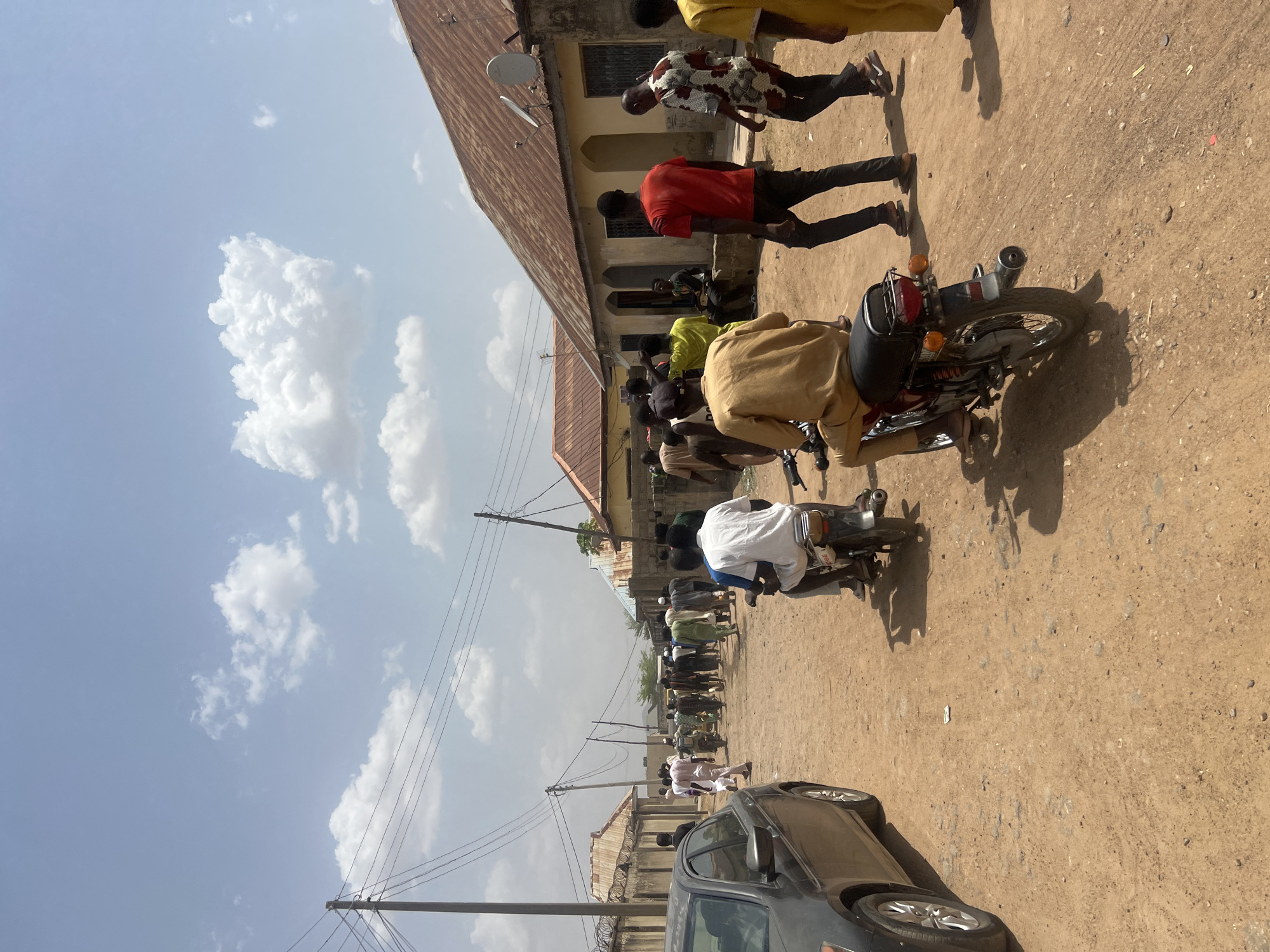 The image captures a vibrant daytime street scene in Minna, Nigeria, depicting everyday activity on an unpaved dirt road. The perspective is slightly elevated, looking down the length of the street.
Numerous people, predominantly men, are scattered across the scene. Many are standing or walking, while others are seated on motorcycles. Their attire varies from t-shirts and trousers to traditional loose-fitting garments. Several motorcycles are visible, with individuals riding or sitting on them. In the foreground, partially visible on the right, is a dark-colored car, possibly a sedan. Along the left side of the street, buildings with brown corrugated metal roofs and simple concrete facades line the road, some featuring windows with security grilles. Utility poles with multiple power lines are prominent against the sky, extending down the street.
The activity suggests a regular day on a busy street. People are going about their daily routines, either moving on foot or utilizing motorcycles for transport. The scene appears to be a casual gathering or a typical flow of traffic and pedestrian movement.
Notable details include the bright daylight, indicated by the clear shadows cast by people and vehicles on the dusty ground, suggesting a mid-day to early afternoon setting. The sky is light blue with scattered white cumulus clouds. The ground is a packed dirt road with small stones, typical of unpaved areas. The general architecture and mode of transport align with the provided location context of Minna, Nigeria. Visible text on the rear of one motorcycle includes "YAMAHA" and some faint numbers, possibly "02", near the license plate area. The overall atmosphere appears calm and active.