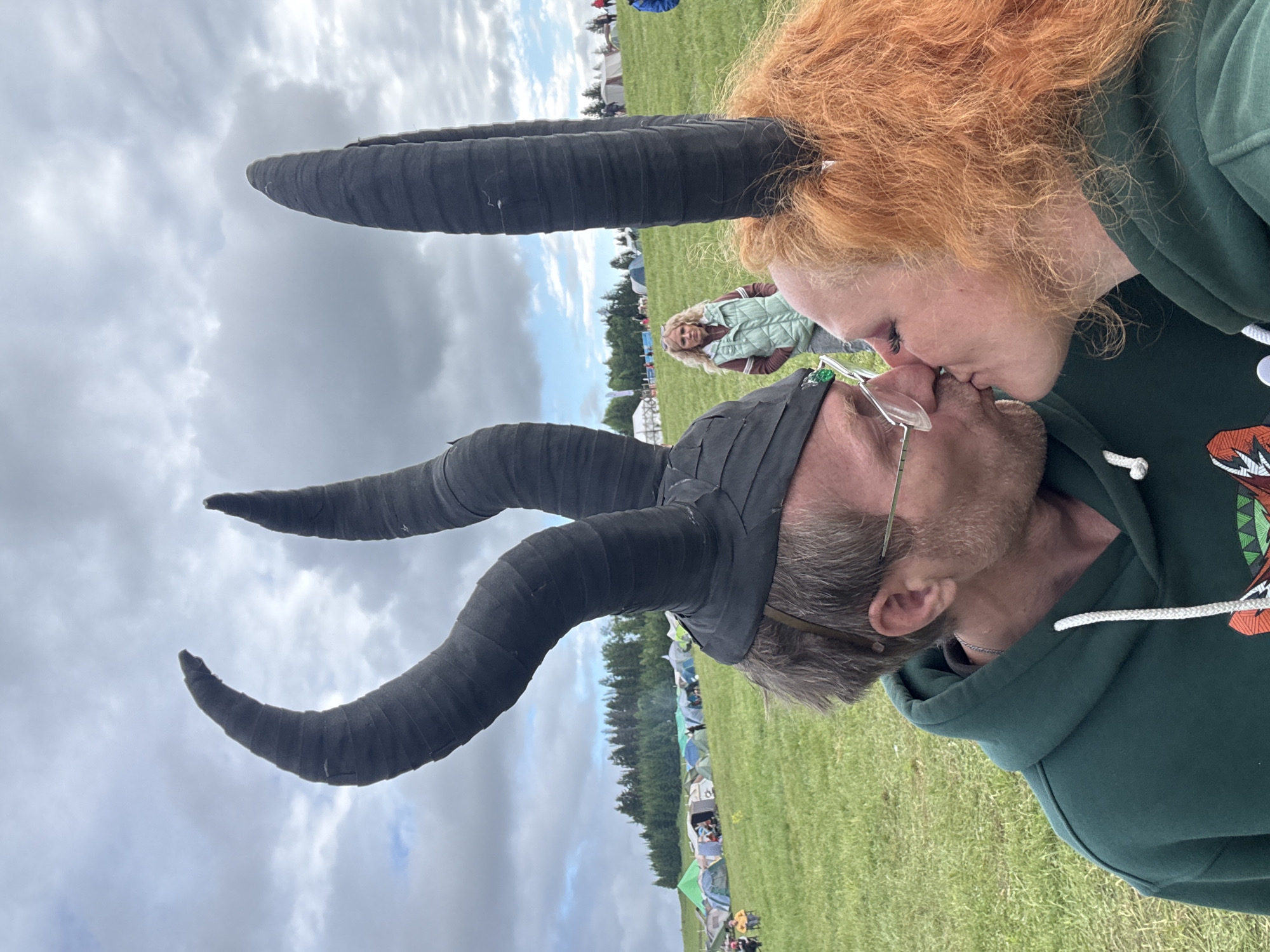 The image depicts an outdoor scene in a large, green grassy field, identified as Bol'shaya Kivara, Russia, beneath an overcast sky. In the immediate foreground, a man and a woman are engaged in a kiss. The man, with short dark hair and wearing glasses, has a dark green hooded sweatshirt and a black headpiece adorned with multiple prominent, spiraling horns. The woman, characterized by vibrant reddish-orange curly hair, also wears a dark green hooded sweatshirt and a black headpiece featuring two large, upward-tapering horns.
In the middle ground, a blonde woman wearing a light mint green quilted vest over a maroon top stands with a neutral expression, positioned between and slightly behind the kissing couple. The background showcases a wide expanse of the field dotted with numerous multi-colored camping tents (including green, blue, and white) extending towards a dense line of evergreen trees that forms the horizon. The overall environment suggests a large outdoor gathering or festival.