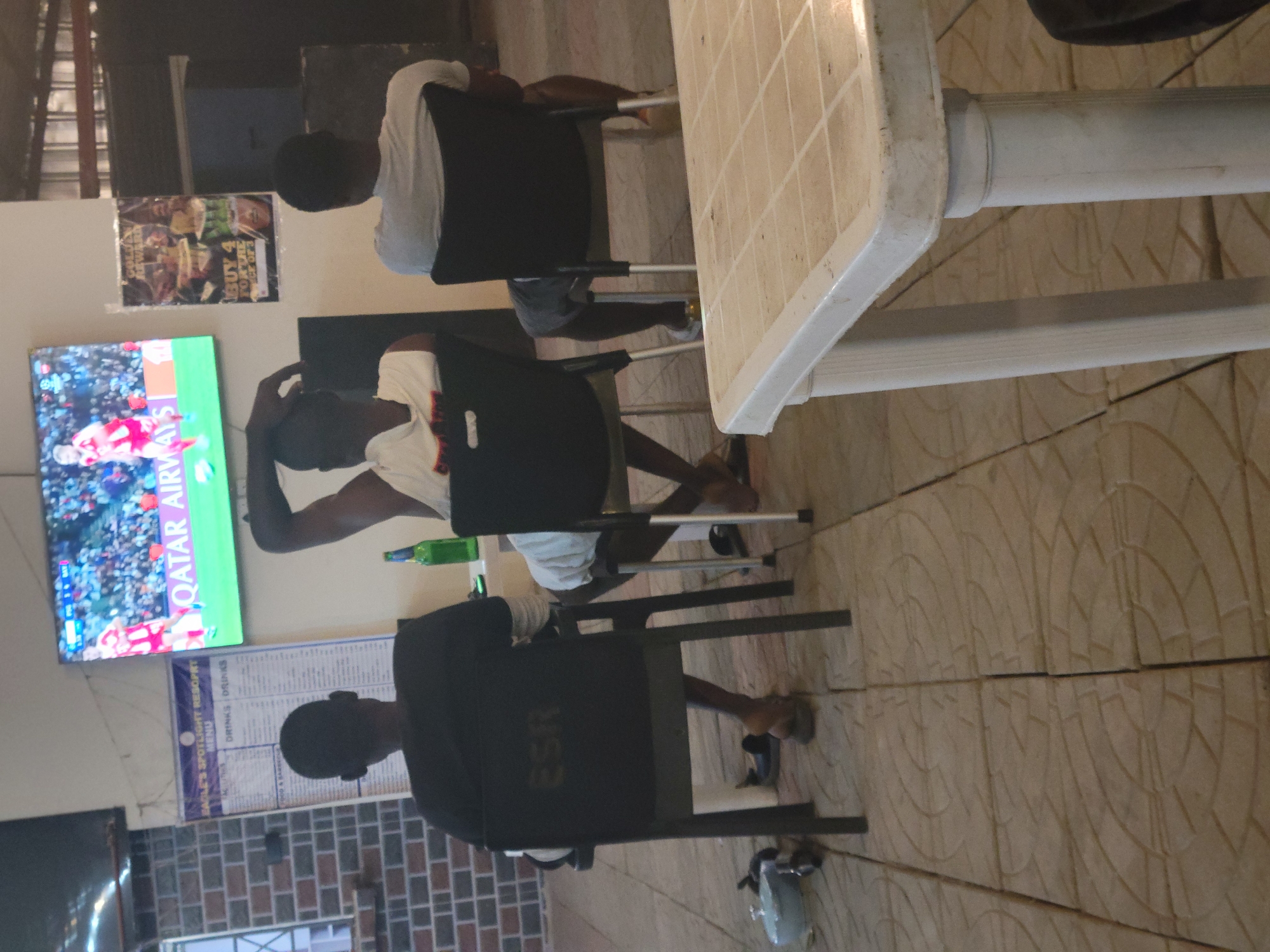 This image captures an indoor scene, likely a commercial establishment such as a sports bar or viewing center, in Ibo, Nigeria. Three men, viewed from behind, are seated on black chairs with metal frames, intently watching a football (soccer) match displayed on a wall-mounted television screen. The TV shows part of a game with players in red and white jerseys, and the text "QATAR AIRWAYS" is prominently visible, suggesting a sponsored broadcast or a major event like the FIFA World Cup.
In the foreground, a white plastic table with a beige tiled pattern covers the right side of the frame. A green bottle stands on the table closest to the central figure. The floor is covered with large, light-colored tiles featuring an intricate circular design. The walls exhibit different finishes: a section on the left displays a reddish-brown brick pattern, while another section is plain, off-white.
Visible text includes "QATAR AIRWAYS" on the television. On a white poster mounted on the wall, partially obscured, the words "LE'S SPOTLIGHT RESORT MENU" and "DRINKS DRINKS" are discernible, indicating a menu board. Another poster on the wall advertises a promotion with text like "BUY 4 FOR THE PRICE OF 3" and "REWARD". The initials "ESR" are visible on the backrest of one of the chairs. The lighting suggests an indoor setting, possibly during the evening. The scene depicts a casual gathering of patrons enjoying a sports broadcast.