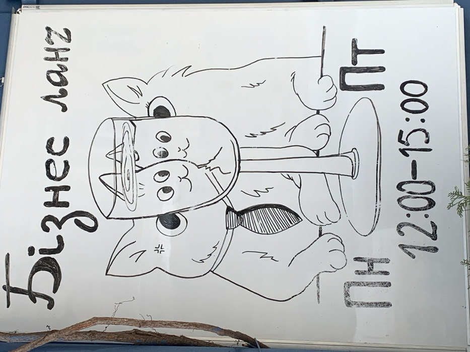 A white rectangular whiteboard, serving as an advertising sign, is affixed to an exterior blue surface in Kyiv, Ukraine. A dry, bare branch partially covers the left edge of the board. The sign features a hand-drawn, black-and-white cartoon illustration of two large anthropomorphic cats flanking a stemmed drinking glass. The cat on the left wears a striped necktie and exhibits a small "angry" symbol near its left eye. The cat on the right is positioned to the right of the glass. Inside the glass, two smaller, smiling cat faces are depicted emerging from a liquid surface with visible ripples. Above the illustration, the Cyrillic text "Бізнес ланч" (Business lunch) is written. Below the illustration, the Cyrillic abbreviations "ПН" (Monday) on the left and "ПТ" (Friday) on the right are displayed, with the time "12:00-15:00" centered between them. Green foliage is visible at the bottom of the frame.
