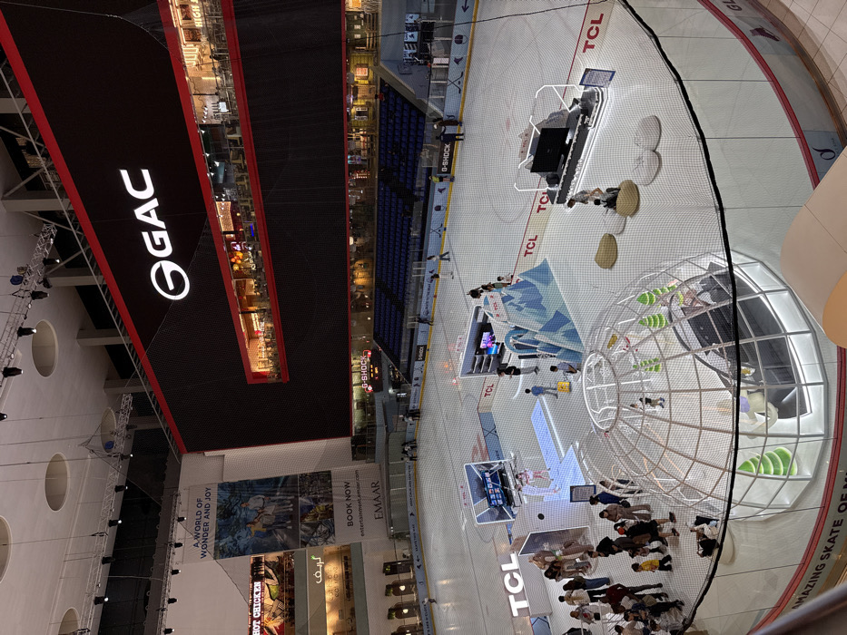 The image captures a wide, high-angle view of a bustling indoor entertainment complex, likely a large shopping mall, looking down onto an ice rink and surrounding structures.
In the upper-left and central part of the image, the multi-story architecture of the mall is visible, featuring several levels of shops or restaurants with warm interior lighting. A very large black banner or screen, edged in red, spans across the upper section, prominently displaying a white "GAC" logo. Below this, various other banners are visible, including one that reads "A WORLD OF WONDER AND JOY" and another with "BOOK NOW entertainment.emaar.com EMAAR". Further down, smaller banners feature "HOT CHICKEN" and the Arabic word "گریف" (Griff). Lighting rigs and ceiling structures are also discernible.
The main activity unfolds on a large ice rink that occupies the lower-right portion of the frame. The rink surface is light gray or white with red and blue lines, characteristic of an ice hockey rink. Several "TCL" logos are clearly visible on the ice. Along one side of the rink, there's a section of blue bleacher-style seating with "G-SHOCK" branding.
Set up on the ice are various interactive exhibits and structures. Two rectangular booths feature large screens and are surrounded by people. Another installation appears to be an abstract, light blue and white structure, possibly resembling ice formations or mountains. A particularly striking feature is a large, spherical, cage-like structure made of white frames, with a clear, domed section. Inside this sphere, green, leaf-like decorative elements or seating are visible, along with people.
Numerous people are scattered across the ice rink area, mostly standing and engaging with the exhibits or observing the scene. They appear to be casually dressed. A black protective net runs across the image, separating the elevated viewing area from the rink below. Partially visible text along the border of the rink reads "AMAZING SKATE OF".
The scene suggests a public event, possibly a product launch, exhibition, or family entertainment program taking place on the ice rink, sponsored by various brands like GAC, TCL, G-SHOCK, and EMAAR. The image is well-lit, indicating an indoor setting, and captured from a high vantage point, offering a broad perspective of the vibrant activity below.