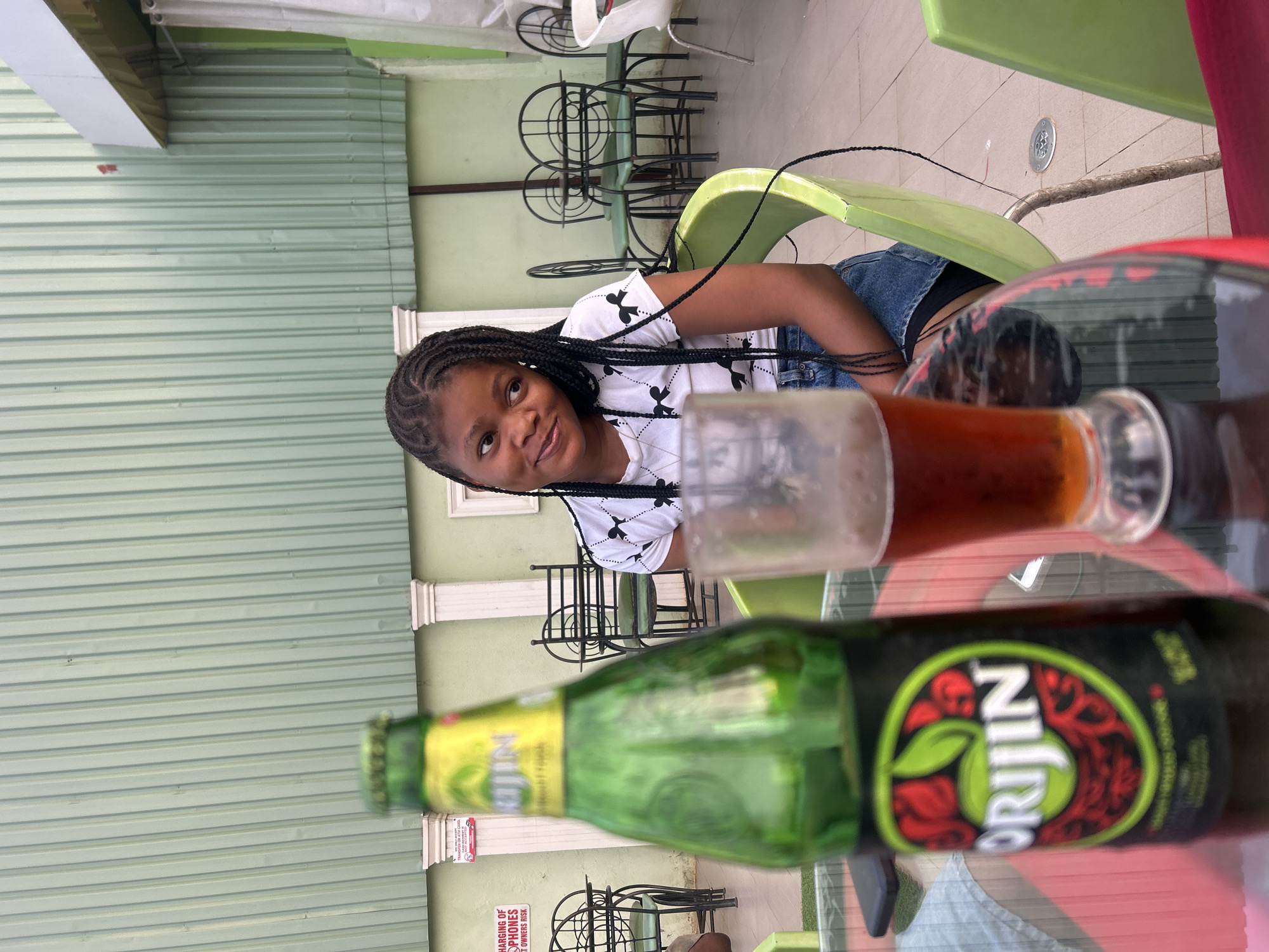 A woman is sitting at a table with alcoholic beverages in Unknown, Unknown. The woman has dark skin, is wearing a white t-shirt with a black pattern, and blue denim shorts. Her hair is braided. On the table, there is a green bottle of "Orijin" and a glass of a dark brown beverage. Behind the woman are green chairs and a light green building. The scene appears to be outdoors, potentially at a restaurant or patio.