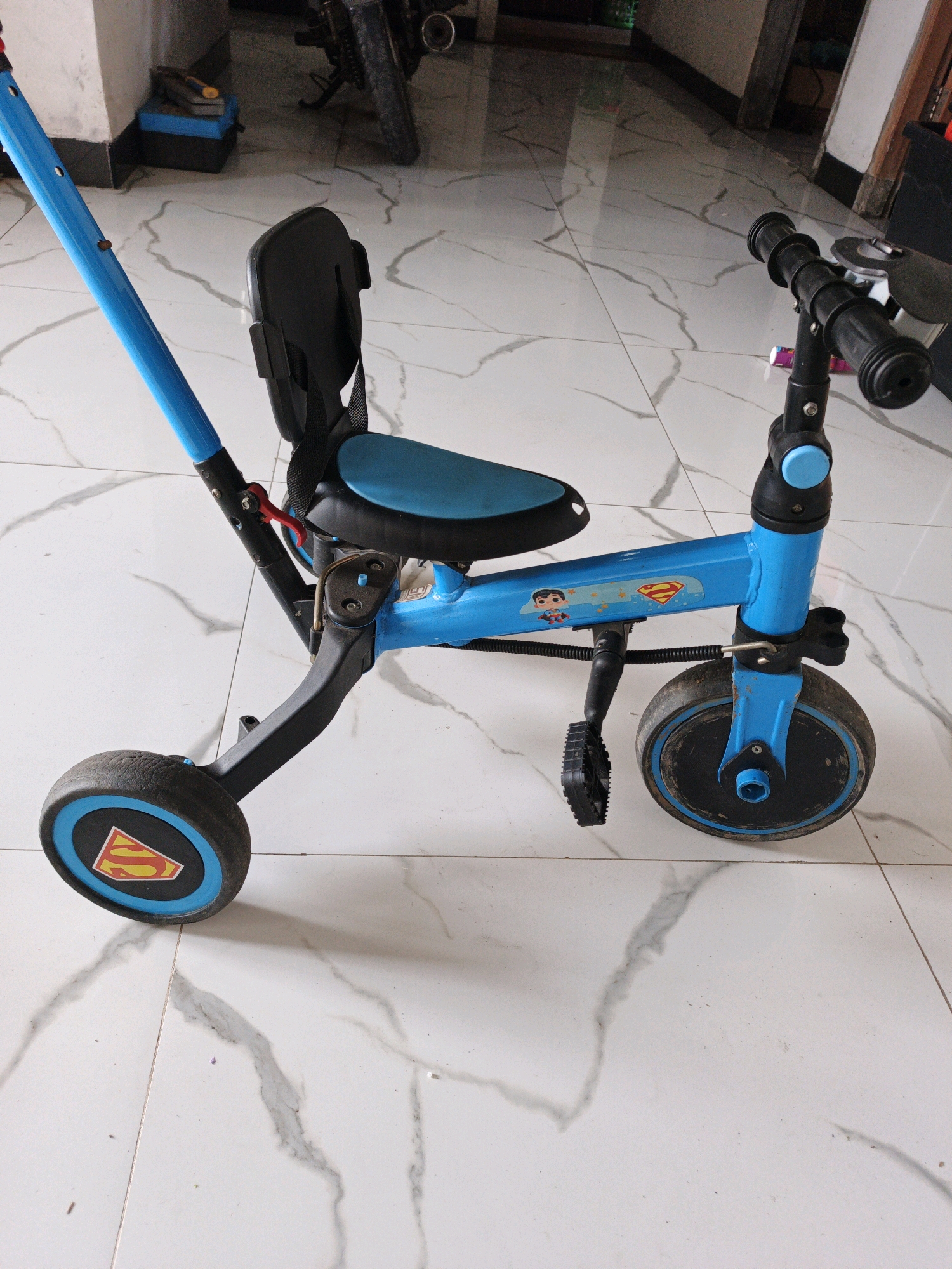 The image shows a blue and black children's tricycle with a push handle, parked on a tiled floor. The tricycle features a blue seat, black handlebars with grips, and a single front wheel with a black tire and a blue rim. The front wheel also has a Superman logo sticker. There are two rear wheels, one of which is visible and has a similar blue rim and a black tire. The tricycle has pedals and a black frame. In the background, a portion of a motorcycle and some boxes are visible. The tiled floor has a white and gray marble pattern. The location is specified as Jatipasar, Indonesia.