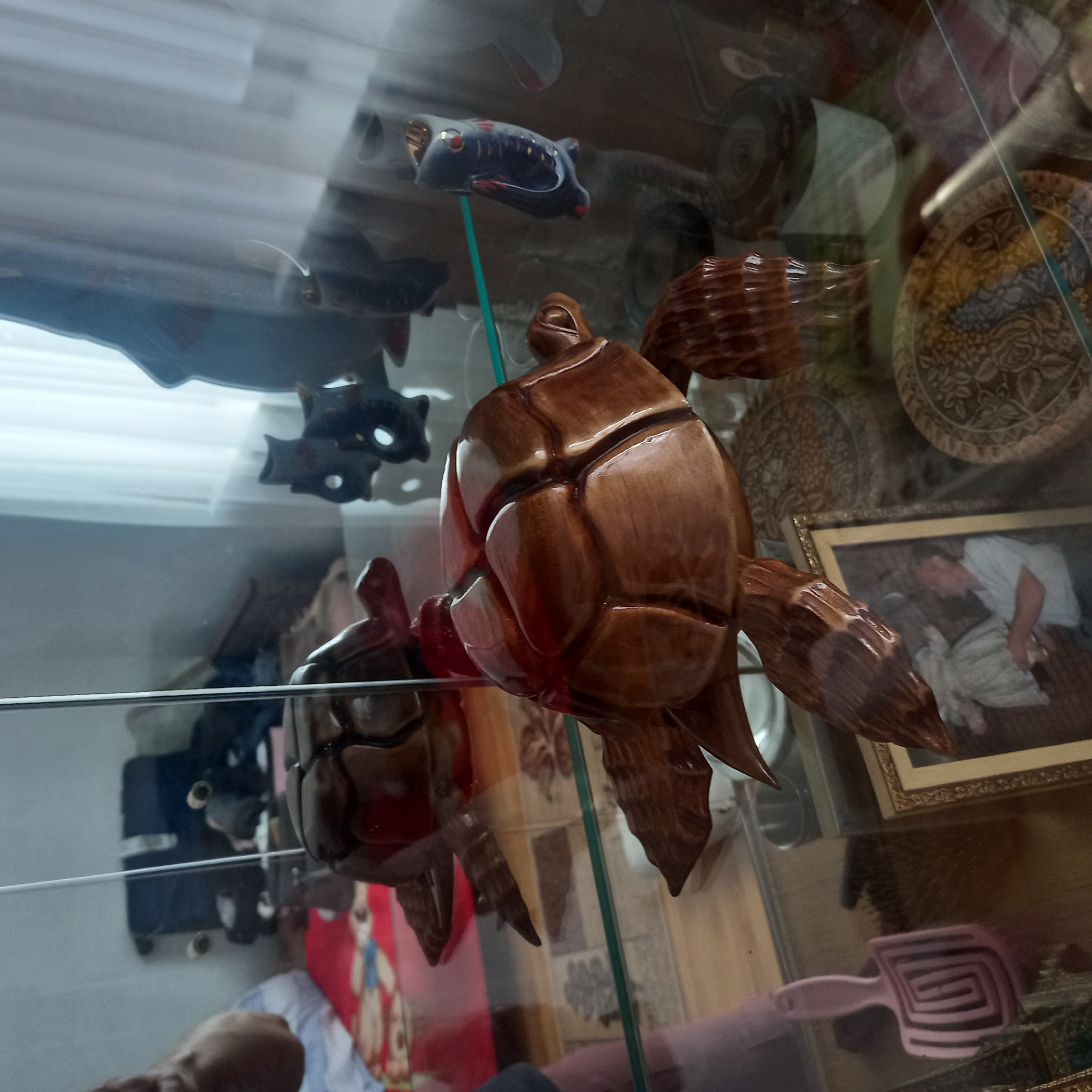 The image shows an interior scene, likely a room in a residence in Cepeleuți, Moldova, as evidenced by context clues. The primary focus is a collection of decorative items displayed within a glass-fronted cabinet. In the foreground, a highly polished, wooden sculpture of a turtle is prominently positioned. This sculpture is intricately carved, with visible details on its shell and limbs. Behind it, and slightly to the left, another similar wooden turtle sculpture is partially visible, suggesting a matching pair or a thematic display.
Further into the cabinet, various other decorative objects can be observed. To the right of the foreground turtle, a small, blue ceramic figurine resembling a fish with red accents is visible. In the background, several other dark-colored ceramic or wooden figurines, possibly owls or other animals, are present. A large, dark, silhouette-like object, possibly a statue or a large decorative piece, is also in the background, to the far right, partially obscured by reflections.
Below the cabinet, on a surface that appears to be a shelf or tabletop, a framed photograph is visible. The photograph depicts a man holding a child, both in light-colored clothing. To the left of the photograph, a pink, comb-like object with a spiral pattern is lying down. Several decorative plates or discs with intricate patterns are also visible on the shelves, adding to the overall display of collected items. The environment suggests a home setting where decorative objects are carefully arranged and displayed, possibly for aesthetic purposes or as part of a personal collection. The lighting suggests an indoor setting, possibly with natural light from a window, indicated by the white curtain visible in the upper right portion of the image. Reflections on the glass doors of the cabinet are present, indicating the presence of a viewer or camera positioned in front of the display.