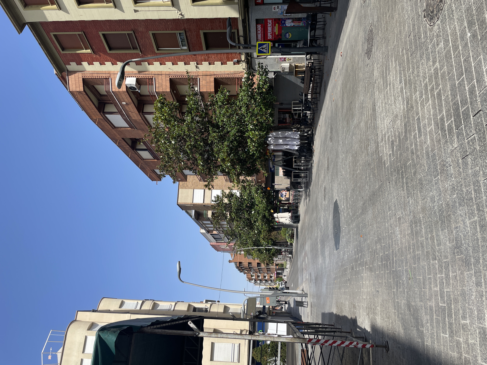 This is a photograph of a street scene in Arnedo, Spain. The image is taken from a low angle, looking up at buildings and the sky. The street is paved with cobblestones and lined with buildings on either side. Some buildings are made of red brick, while others are lighter in color. There are trees planted along the sidewalk, and outdoor seating for cafes or restaurants is visible, with chairs and tables stacked.
In the foreground, there are some metal structures with red and white striped tape, possibly part of a construction or event setup. Further down the street, there are more trees and buildings, and a street lamp is visible. The sky is clear and blue, suggesting a sunny day. The sun is casting shadows on the street, indicating it is likely daytime. There are no people clearly visible in the image, and the overall impression is one of a quiet, urban environment.
Visible text includes a sign that appears to read "SUPERMERCAT" and another with a yellow pedestrian crossing symbol. There are also some small signs on poles further down the street, but the text is too small to read. The scene appears to be set in a commercial or residential area, with a mix of architecture and outdoor amenities.