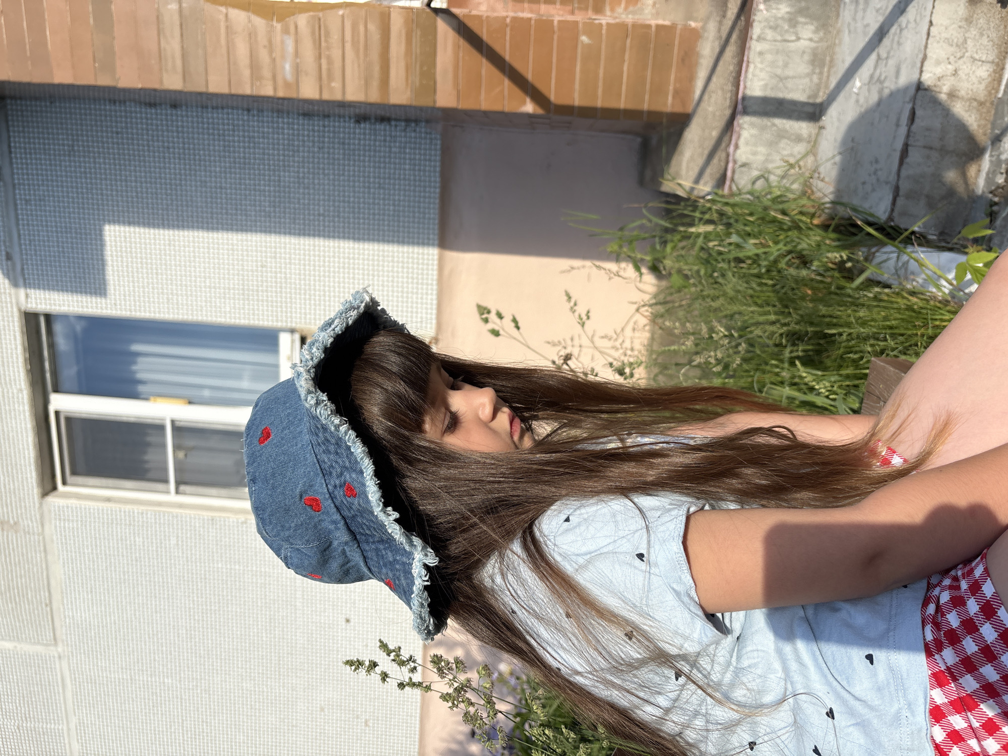 In Chaikovsky, Russia, a young girl with long brown hair, wearing a blue denim bucket hat adorned with red hearts and a light blue t-shirt with a subtle heart pattern, sits outdoors. She is also wearing red and white checkered shorts. She is looking downwards and appears to be in a pensive mood. The background features a building with a light-colored, textured facade and a window. The area is surrounded by green grass and some dried plants. The lighting suggests it is daytime, with shadows cast on the building's surface.