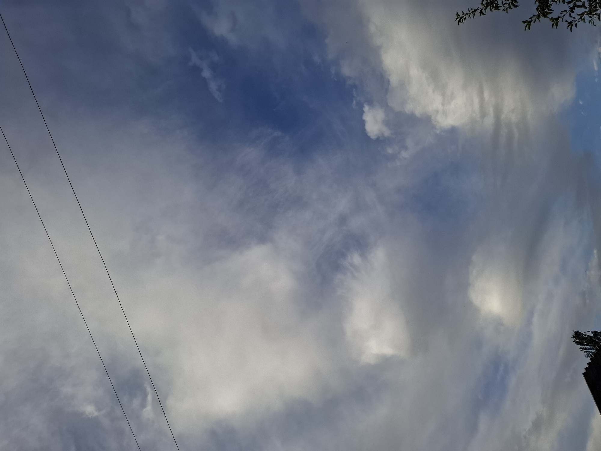 A low-angle shot captures a dynamic sky over Jeti-Ögüz, Kyrgyzstan. Two thin, dark wires stretch diagonally across the frame from the upper left to the lower right. The sky is a blend of deep blue and grey, with varying cloud formations. Wispy, thin clouds are interspersed with larger, brighter, and denser clouds that appear to be closer. The overall lighting suggests it is daytime, possibly late afternoon, with uneven illumination across the cloudscape. The bottom of the image shows a sliver of dark, angled roofline, and the upper portion of a dark-branched tree with green leaves on the right side.