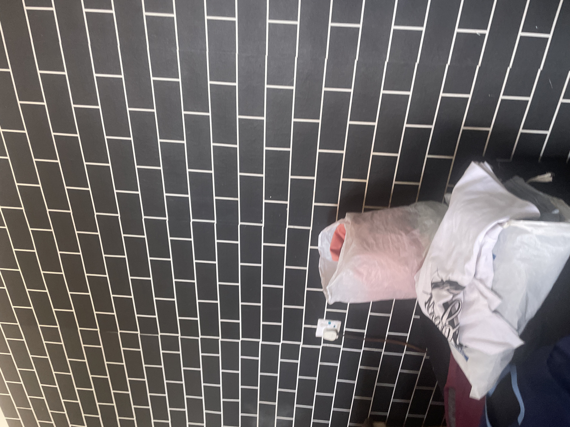 A partially visible interior scene in Jauro Yinu, Nigeria, shows a dark gray brick-patterned wall with white grout lines. To the right of the wall, a large, translucent white plastic bag is filled with a reddish-orange item. Below the bag, stacked clothing items are visible. A white t-shirt with a black graphic design is on top, and other white and dark-colored garments are underneath. To the left of the bag, a white electrical outlet with a plug is mounted on the wall. The scene is lit, suggesting it is daytime or a well-lit interior space.