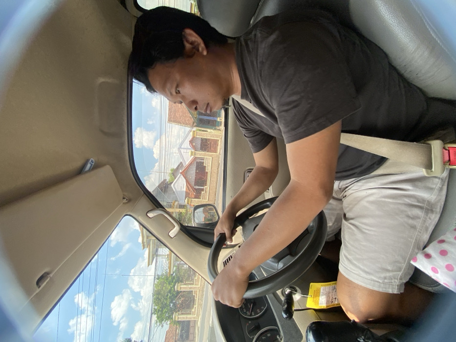 A man wearing a dark shirt and khaki shorts is driving a car. He has a seatbelt fastened. Outside the window, houses and trees are visible under a blue sky with scattered clouds. The sun visor is flipped down on the driver's side. A tag is hanging from the car key in the ignition.