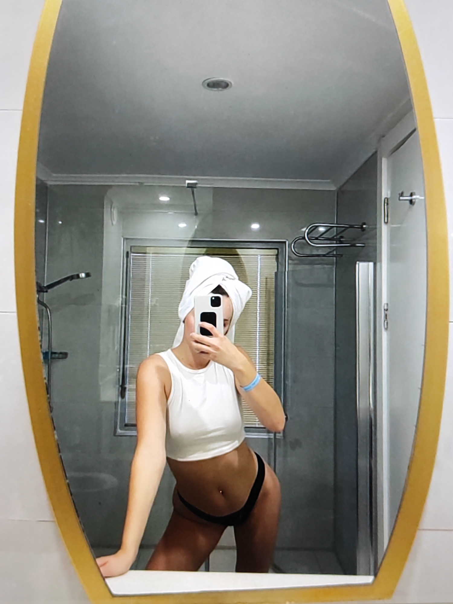 A woman with a towel on her head, wearing a white crop top and black bikini bottoms, takes a selfie in a bathroom mirror. She is leaning on the vanity, and her right arm is extended to hold the phone. A light blue wristband is visible on her left wrist. The bathroom appears to be in a hotel room, with a shower stall, a towel rack, and a door visible in the reflection. The mirror has a gold-colored, oval frame. The image does not provide any specific indicators to place it within Fergana City, Uzbekistan.