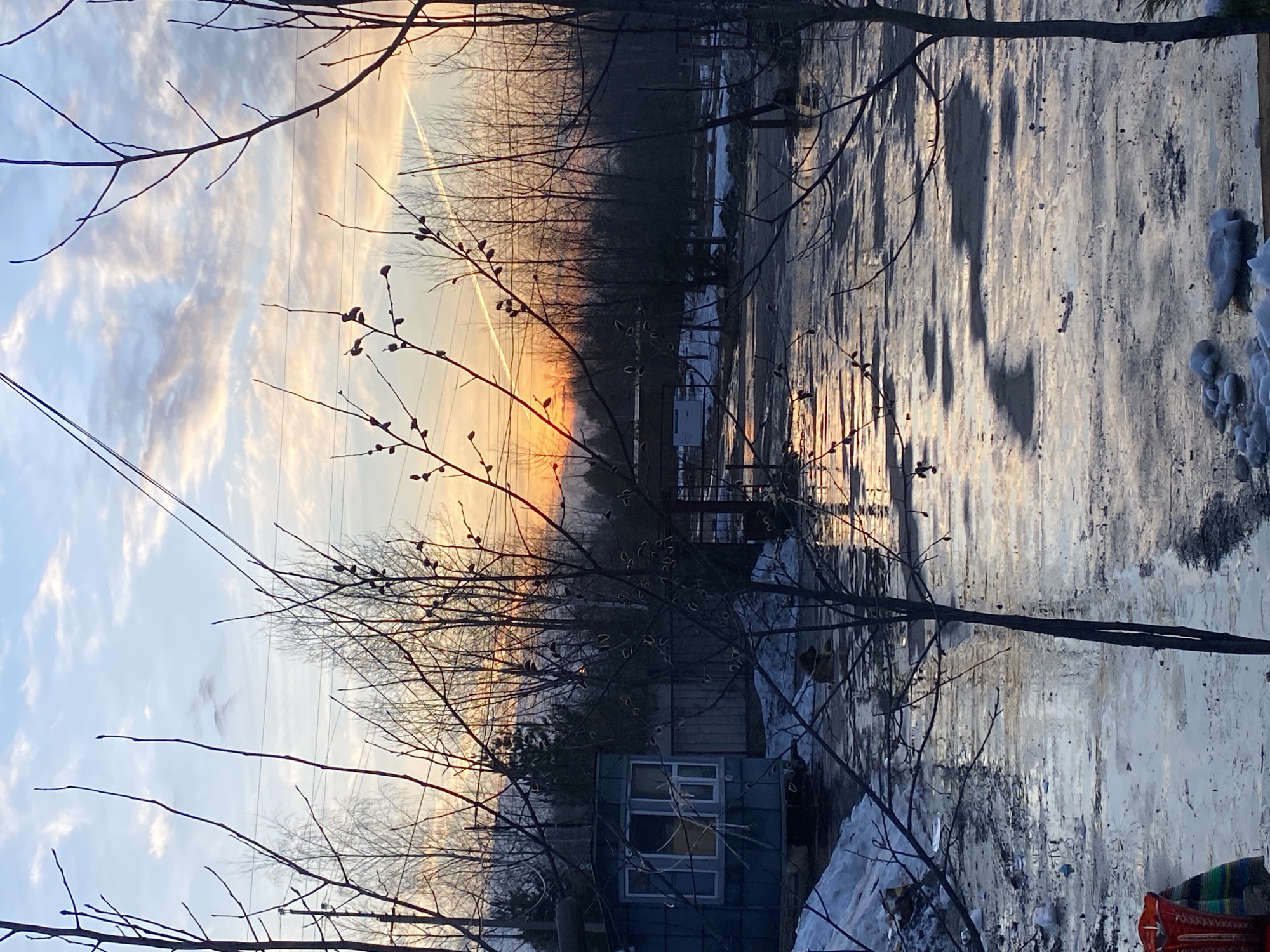 The image depicts a landscape in Ol'khovskoye, Russia, likely taken during winter or early spring due to the presence of snow. The foreground features bare trees with budding branches, obscuring a blue shed-like structure with windows. In the middle ground, there is a road or yard covered with ice and patches of snow. The background reveals a line of bare trees silhouetted against a sunrise or sunset with a distinct orange glow. Power lines are visible in the sky. The sky itself is filled with clouds. A contrail is noticeable in the upper portion of the sky.