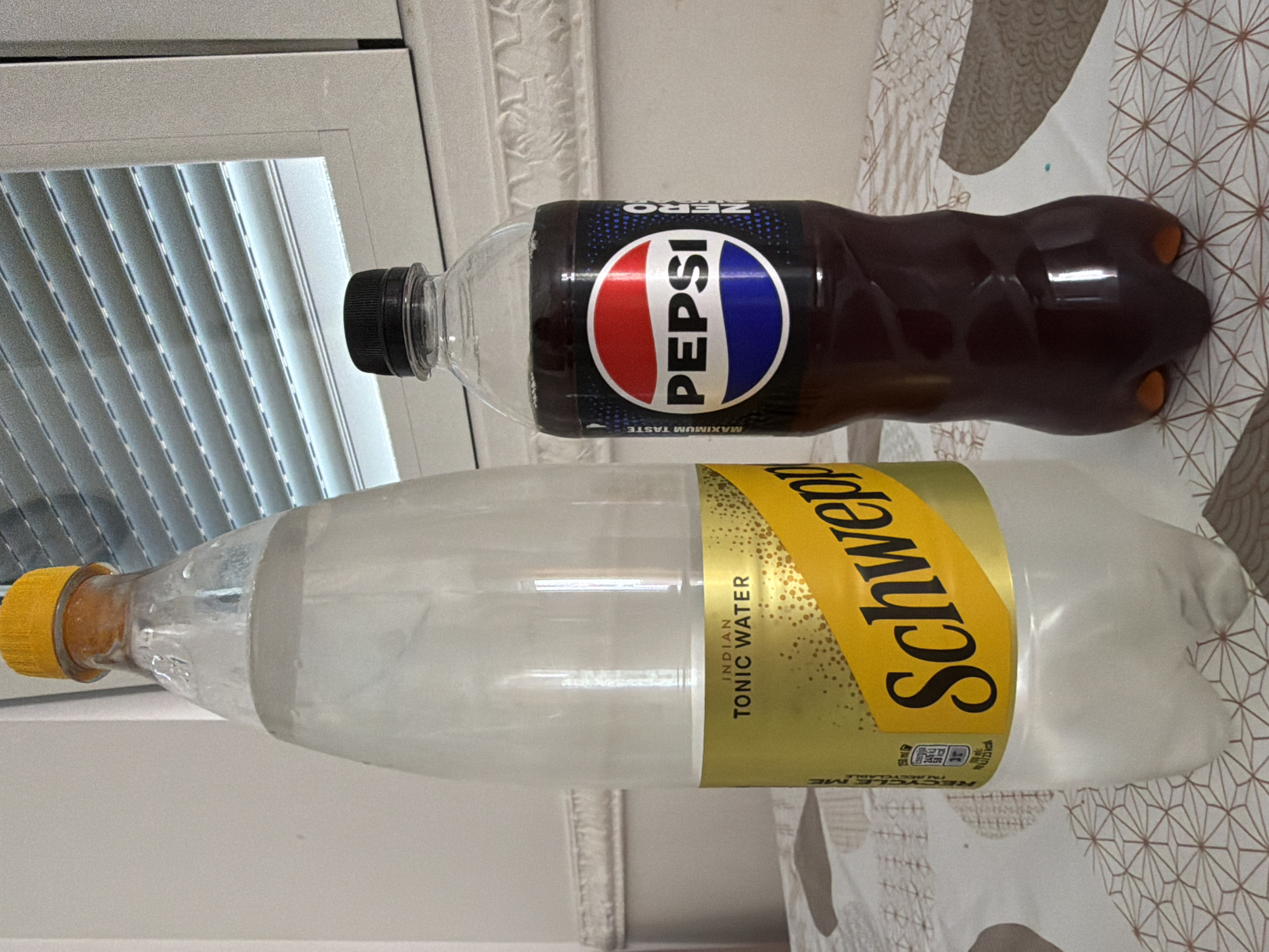 A bottle of Schweppes Indian Tonic Water and a bottle of Pepsi are placed on a table with a patterned tablecloth. The Schweppes bottle is taller and mostly transparent, filled with a clear liquid, and has a yellow label with "INDIAN TONIC WATER" printed on it. The Pepsi bottle is shorter, filled with a dark brown liquid, and has a black label with the iconic Pepsi logo. Both bottles have clear plastic caps. In the background, there is a window with white blinds. The scene appears to be indoors, possibly in a home or hotel room. No people are present in the image. The lighting suggests it is daytime. The specific location in Budva, Montenegro cannot be determined from the image alone.