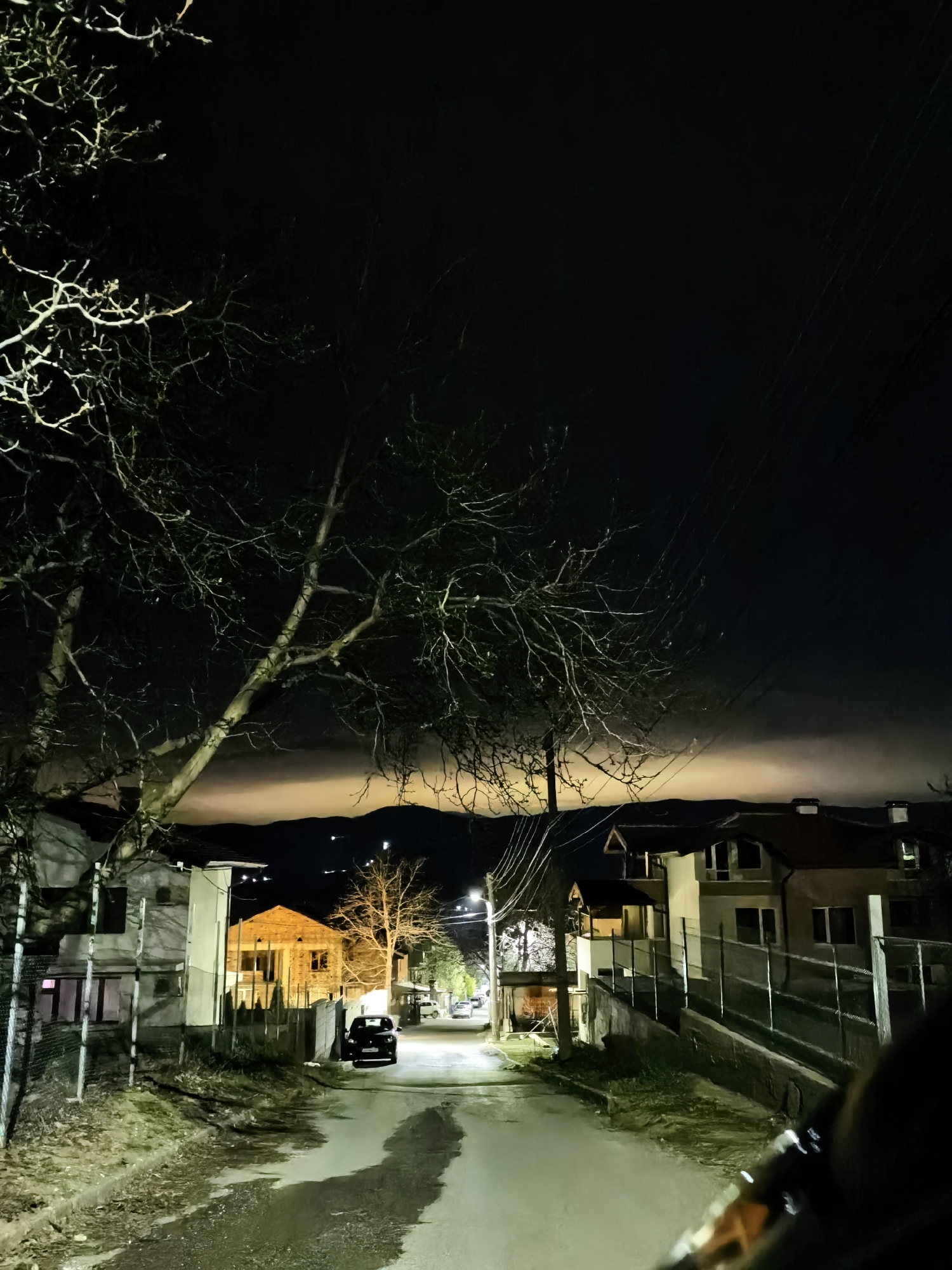 At night, a street in an unknown city is captured. The road is paved, and damp patches create reflections in the light. There are houses on both sides of the street, with fences partially bordering the properties. A black car is parked on the right side of the road, halfway down the street. Bare trees are positioned on the left of the frame. Mountains are vaguely visible in the distance, with a layer of light above them, hinting at either city lights or a fading sunset. Power lines stretch across the sky above the houses.
