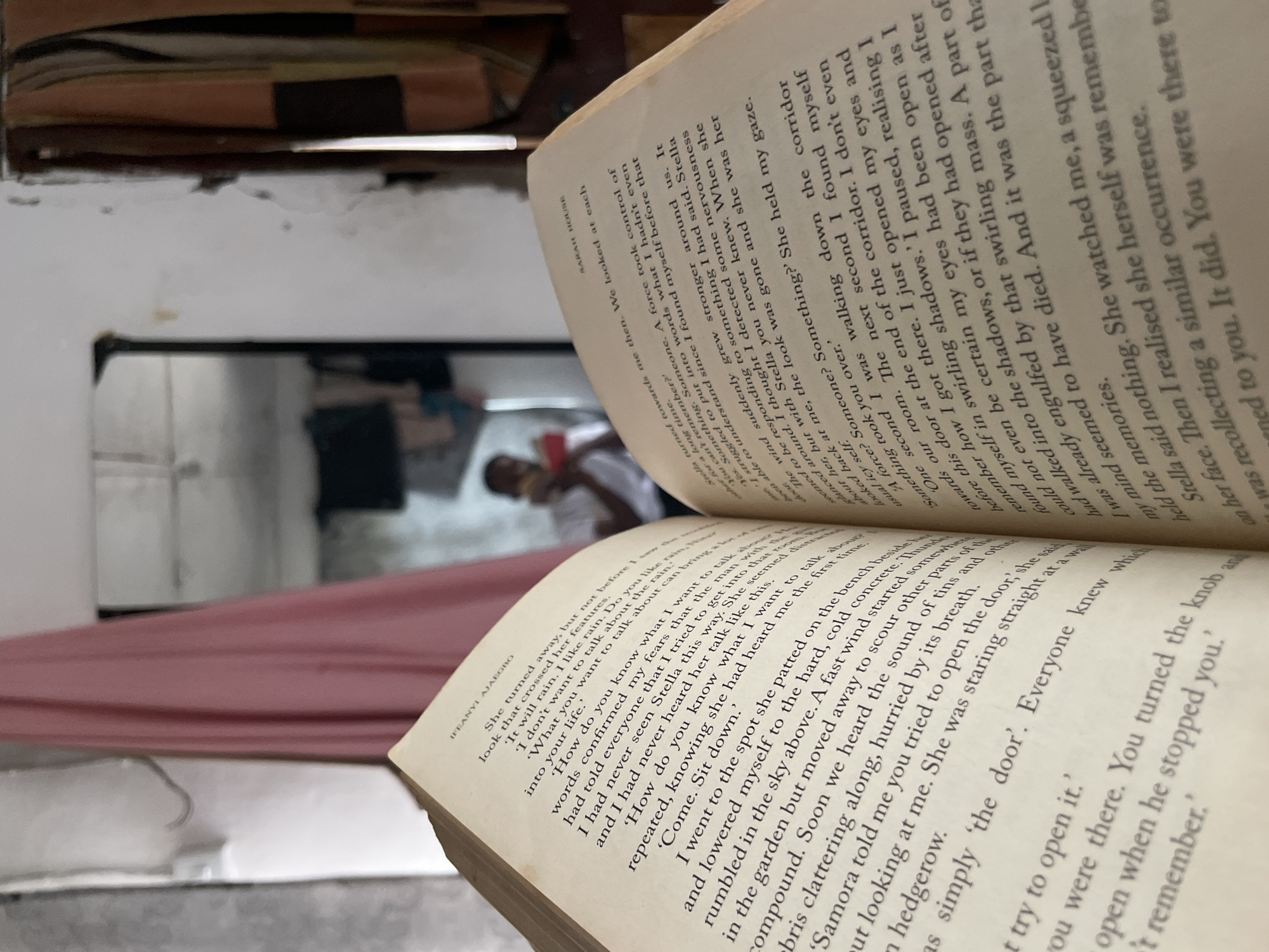 The image is taken through an open book, with text in English visible on the pages. The book obscures much of the background. A person is visible through an opening, seemingly a doorway, in the background. The walls appear to be white and somewhat damaged. The setting is ambiguous, but given that the OCR of the text in the book mentions "IFEANYI AJAEGBO", it could be in Nigeria, specifically Lokoja, as requested, though this cannot be definitively determined from the visual information. The focus is shallow, with the book in the foreground being sharp and the background somewhat blurred.