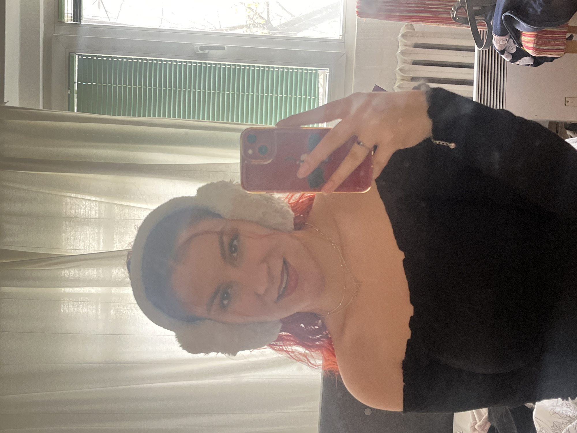 A young adult female is taking a selfie in what appears to be an interior space. She is wearing white earmuffs and a black off-the-shoulder top. She has red hair and is wearing a necklace. She is holding a red smartphone, presumably to take the photo. Behind her is a window with closed blinds and sheer white curtains. A radiator is also visible in the background. There is no information to confirm that this image was taken in Sofia, Bulgaria. Therefore, I cannot provide any details about the location in that city.