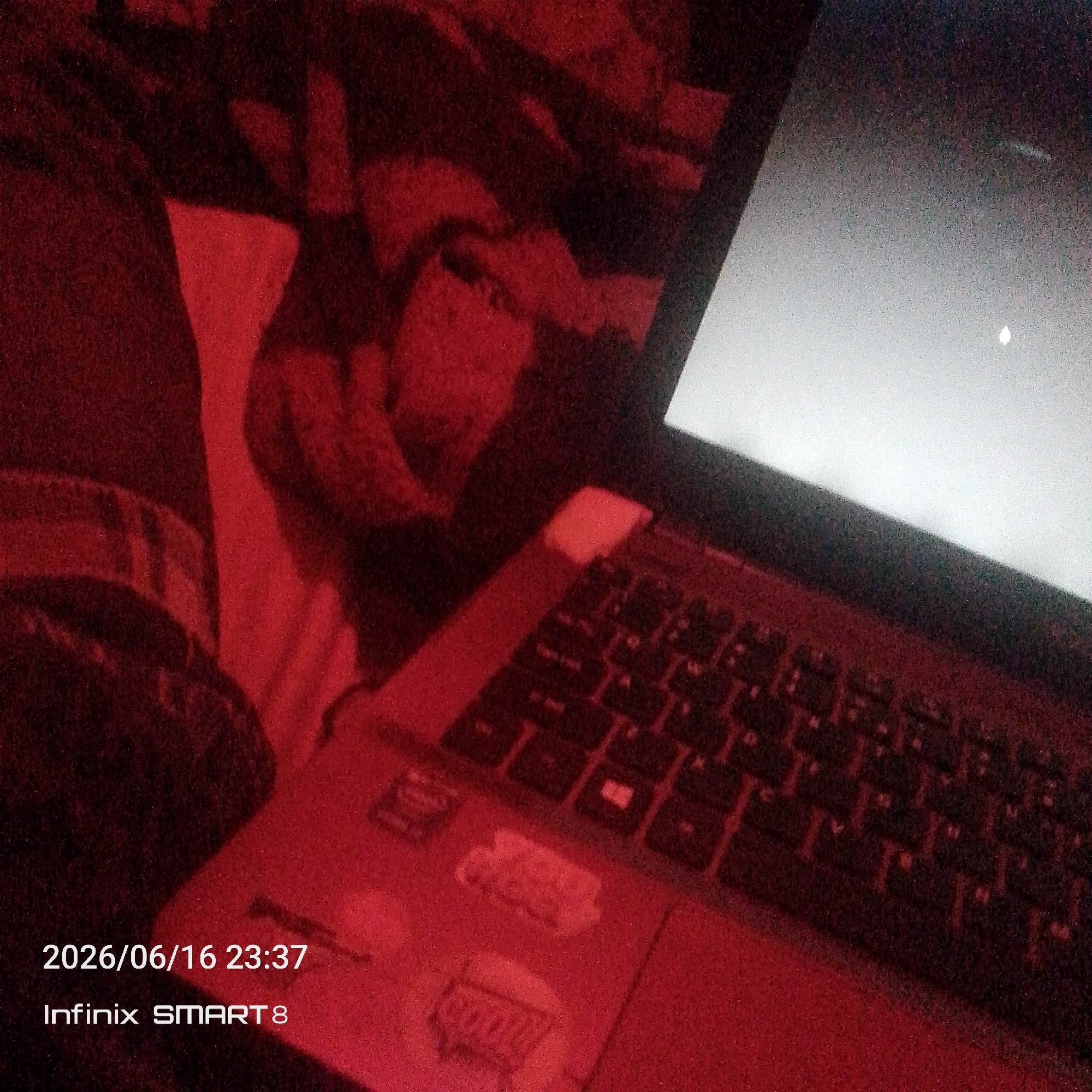 The image is a close-up, dimly lit photograph taken at night, as indicated by the timestamp "2026/06/16 23:37". The scene appears to be indoors, possibly a bedroom or living area, with a strong red cast from an artificial light source. In the foreground, a laptop is open and its screen is illuminated with a bright, uniform light, suggesting it is on. The keyboard of the laptop is visible, with keys illuminated by the same red light. To the left of the laptop, a person's leg, clad in dark fabric, is partially visible. The background is obscured by shadows and the red lighting, but it seems to consist of fabric with a patterned texture. The text "Infinix SMART 8" is overlaid on the bottom left of the image, indicating the model of the device used to capture the photograph. There is no discernible information within the image to suggest a specific location within the city of Jos, Nigeria.