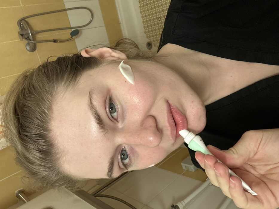 A woman with fair skin applies cream to her lips from a white and mint green tube. Another dab of cream sits on her right cheek. She is angled towards the camera, with her face in focus. The background includes a shower fixture on a yellow tiled wall and a grid patterned floor. The woman wears a black top.