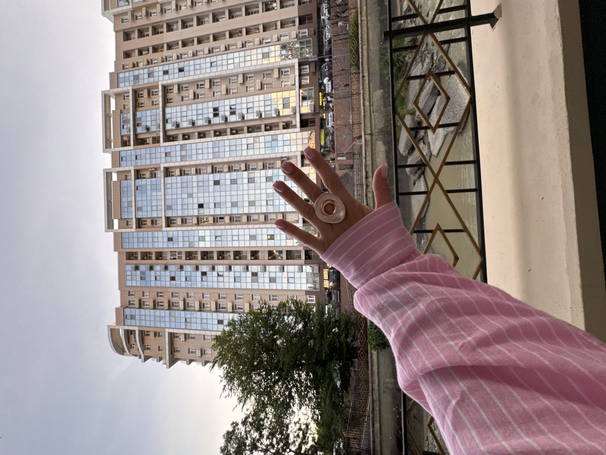 A person's hand, with light pink fingernails and a ring on the index finger, is held up in the foreground, obscuring a portion of a tall, modern apartment building. The building, a light brown color with large glass windows and balconies, dominates the background. Trees are visible to the left of the building. In the mid-ground, a body of water flows between rocky banks, with a decorative black and gold metal railing in front of it. The sky is overcast and light gray. The scene appears to be an urban setting, possibly in Sochi, Russia, given the architectural style of the building and the presence of a water channel that could be a river or canal. The overall atmosphere is casual, with the focus on the hand and the ring as a personal element against the urban landscape.