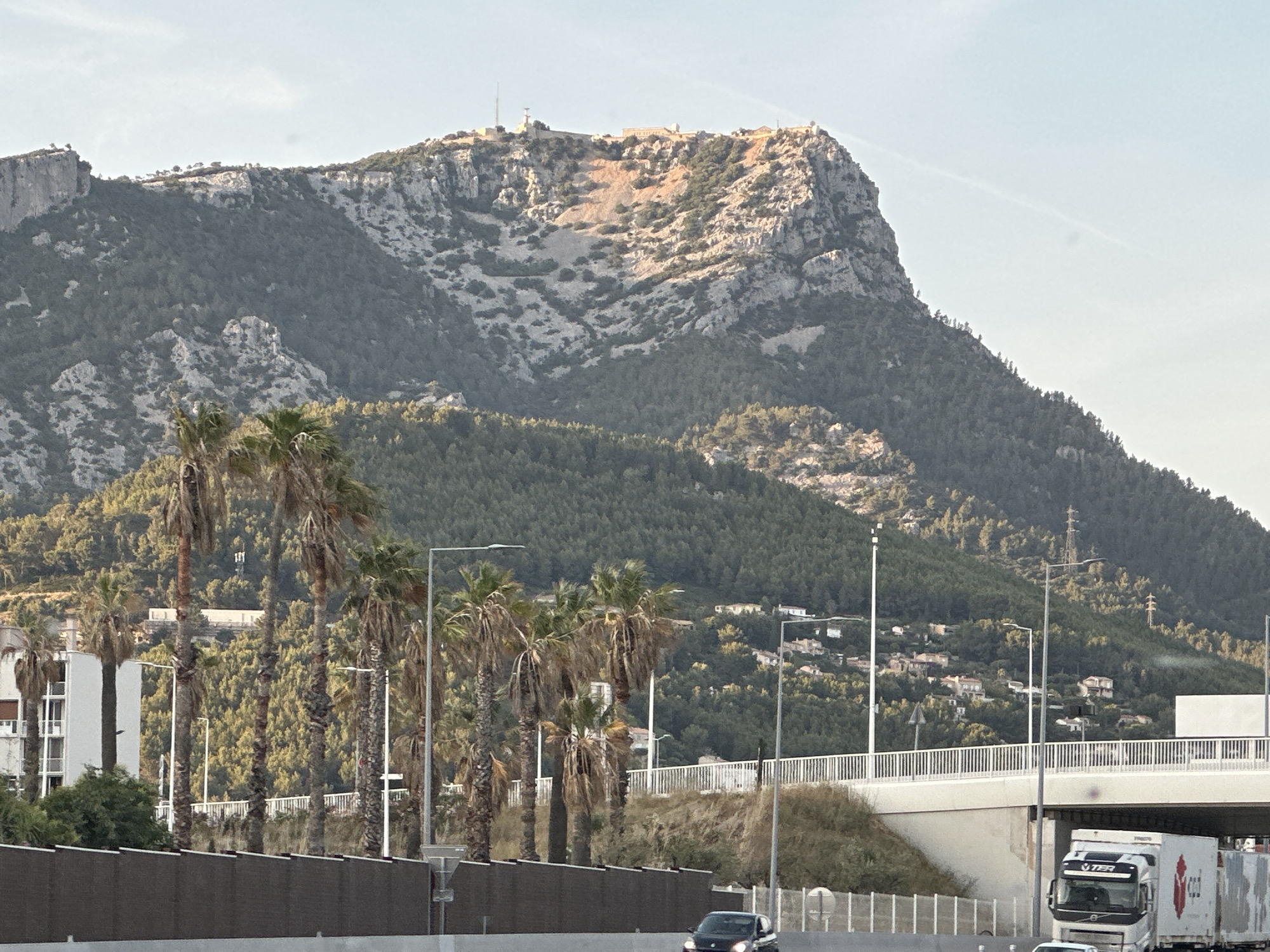 The image shows a wide, mountainous landscape with a city nestled on its slopes, likely in Toulon, France. A prominent rocky mountain dominates the background, its peak adorned with structures, possibly radio towers and buildings. The mountain's slopes are covered in dense green vegetation and sparse rocky outcrops. In the mid-ground, a network of roads and overpasses is visible, with a white truck traveling under one of them. Palm trees line a section of the foreground, adding a tropical element to the scene. The sky is a clear, pale blue with a faint contrail. The overall impression is of a natural landscape integrated with urban infrastructure.