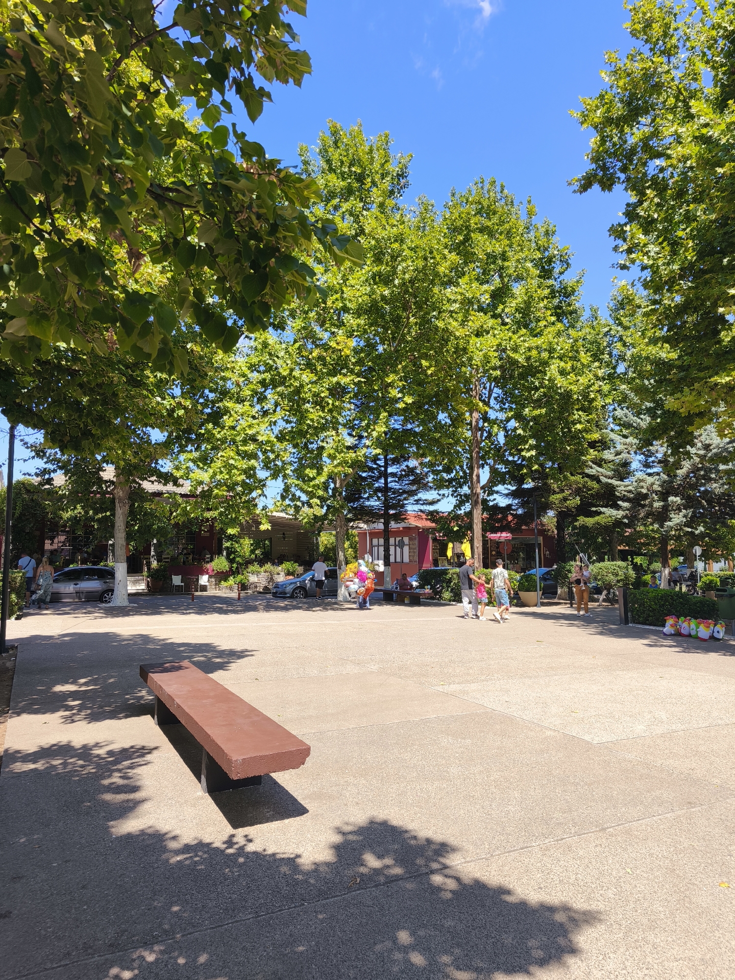 The image depicts an outdoor public space in Prokopi, Greece, bathed in bright sunlight under a clear blue sky. A long, reddish-brown bench is positioned in the foreground, casting a shadow on the paved ground. In the mid-ground, a paved square is populated by a few individuals. To the left, a couple stands near a parked car. Further into the square, a group of people, including children, are walking. Behind them, buildings with red roofs and outdoor seating areas are visible, suggesting a commercial or dining establishment. Tall, leafy green trees line the perimeter of the square, providing dappled shade. To the right, evergreen trees form a backdrop, and a row of colorful, animal-shaped toys are displayed. The overall scene conveys a tranquil and pleasant atmosphere, likely during daytime hours.