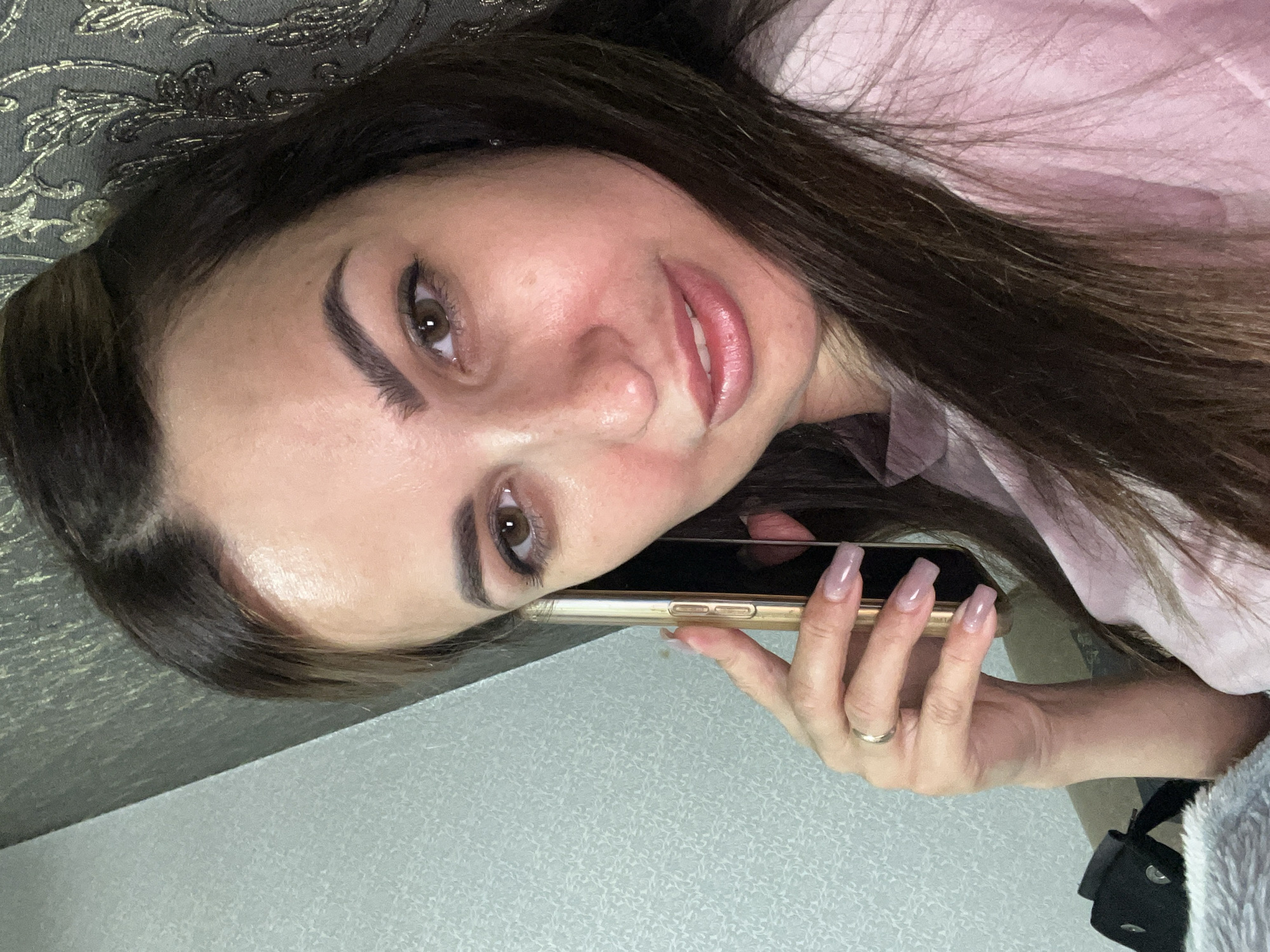 A medium shot shows a woman with dark brown hair holding a golden smartphone to her right ear. She is looking towards the camera with a slight smile. The woman is wearing a light pink top. Her left hand, with long light pink nails and a thin ring on her ring finger, is near her cheek. The background is a textured wall with a floral pattern on the right and a lighter textured pattern on the left. The lighting is soft, casting a slight glow on her face. The scene appears to be indoors, possibly in a bedroom or living room.