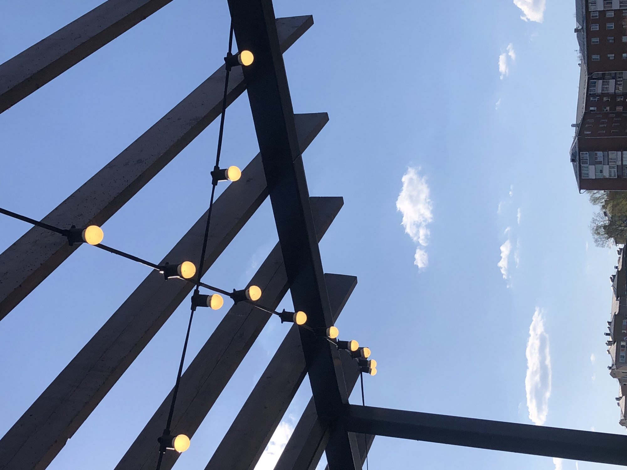 The image shows a low-angle view of a wooden pergola structure with string lights against a blue sky with scattered white clouds. The pergola consists of dark-colored wooden beams arranged in a series of angled lines. String lights with round, yellow bulbs are attached to the beams. In the upper right of the image, the top portion of a brick building is visible. The photograph's perspective is looking upwards. There is no information in the image to indicate that this scene is in Votkinsk, Russia.