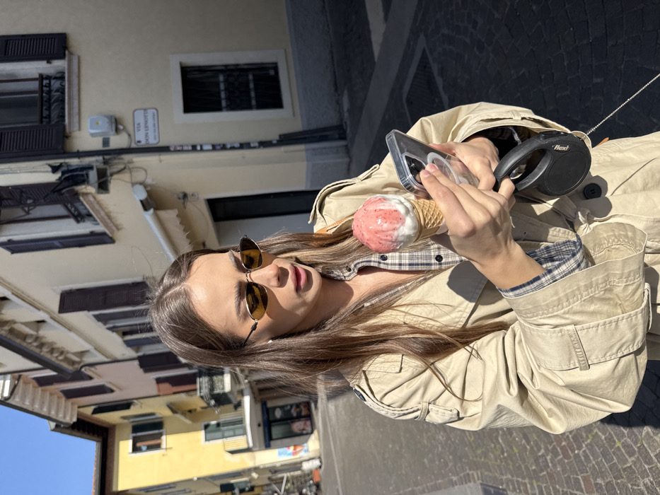 A young adult female stands on Via Don Lenotti in Peschiera del Garda, Italy, under direct sunlight. She has long, light brown hair and wears small, oval-shaped sunglasses with yellow lenses, a light beige trench coat, and a plaid shirt. Her mouth is slightly ajar. In her left hand, she holds a waffle cone containing two scoops of ice cream: one white/cream and one pink/red with red streaks. In her right hand, she holds a clear-cased smartphone, seemingly viewing its screen. A black retractable "Flexi" brand dog leash is attached to or held by her right hand. The background features light-colored, multi-story European buildings with dark window shutters. A street sign on the building to the right reads "VIA DON LENOTTI." The street is paved with dark cobblestones.