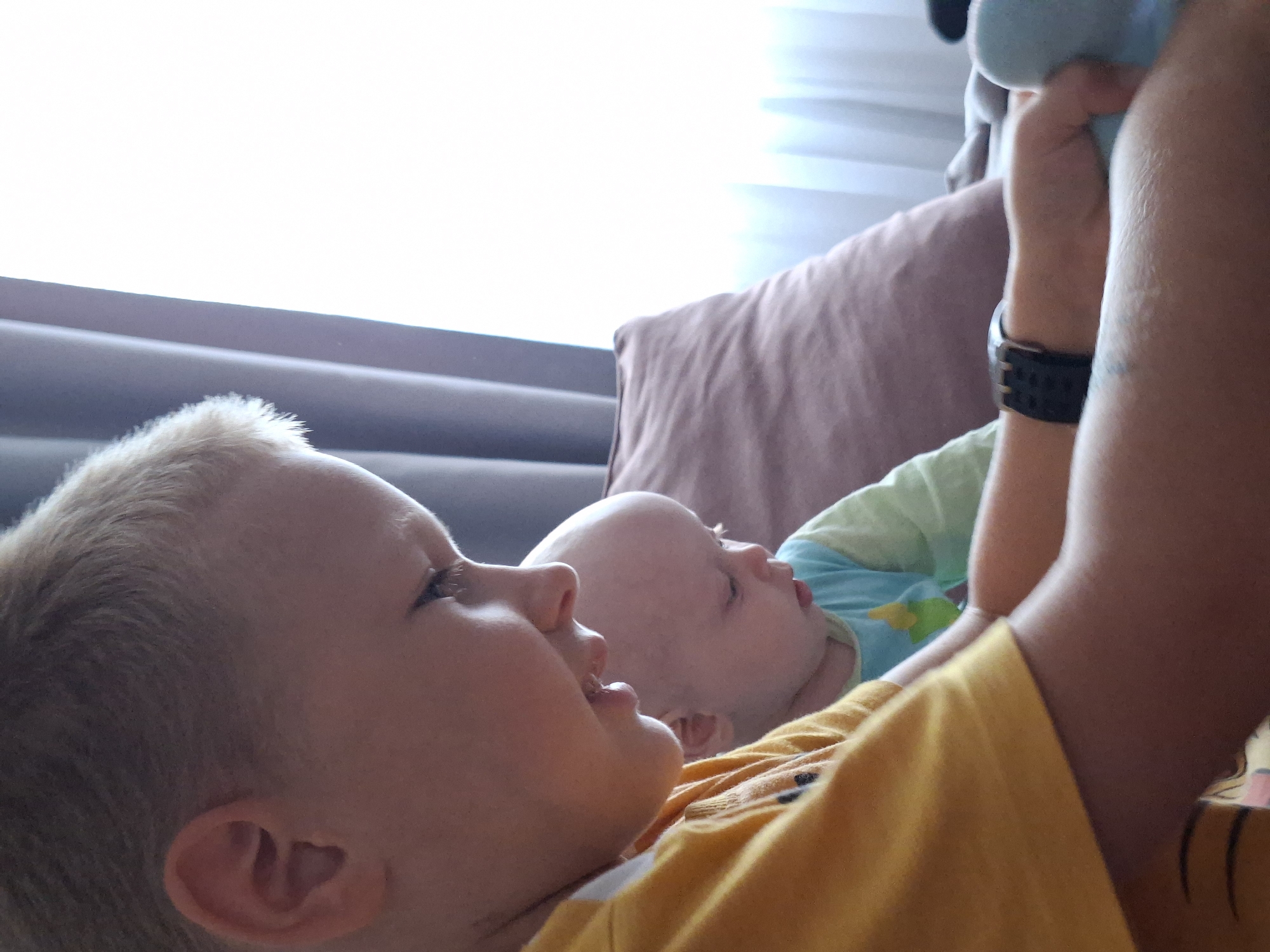 An over-the-shoulder, close-up shot focuses on two young boys engaged in an activity indoors, likely at home. The boy in the foreground, with fair hair, wears a bright yellow t-shirt. His profile is visible as he looks down intently, his mouth slightly open. He appears to be interacting with something or someone below and to his right, his hands are visible and he is wearing a black watch on his left wrist. Behind him, a much younger child, bald with light skin, is also looking downwards, dressed in a turquoise and light green shirt. The background shows a brown textured pillow, grey curtains, and bright daylight filtering through a window. The scene suggests a moment of shared attention or play between the two siblings.