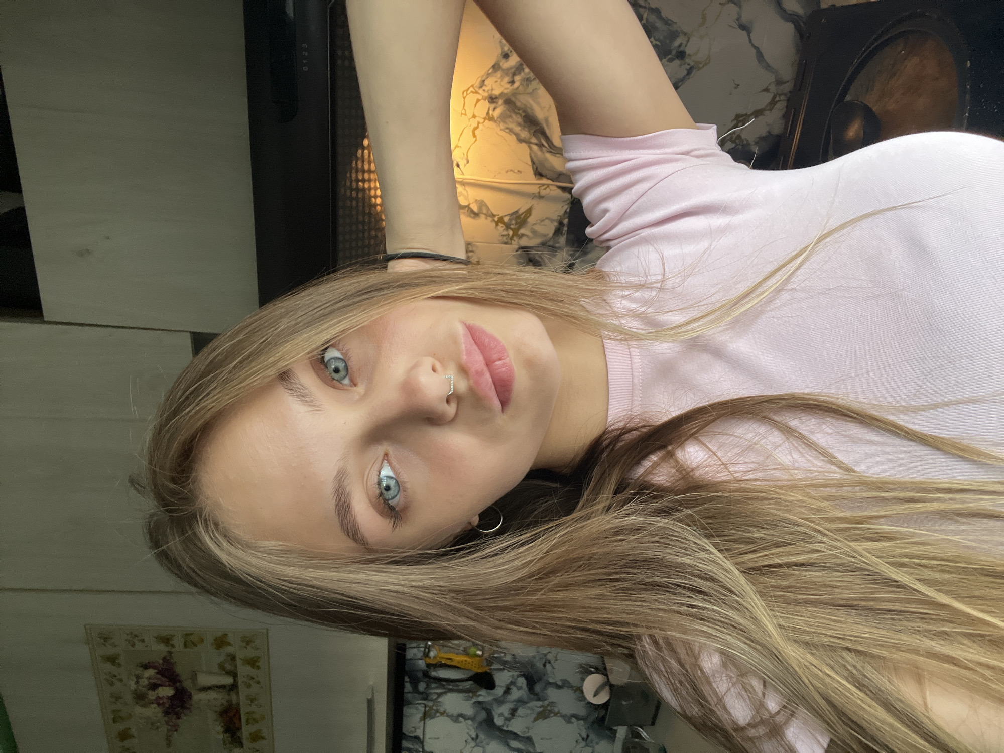 A young woman with light blonde hair and blue eyes is looking at the camera. She is wearing a pink t-shirt. She has a piercing in her nose and an earring. The background includes a kitchen with marble countertops, a black range hood, and cabinets. There is a framed floral picture on the wall. The location is potentially Фоки, Russia.