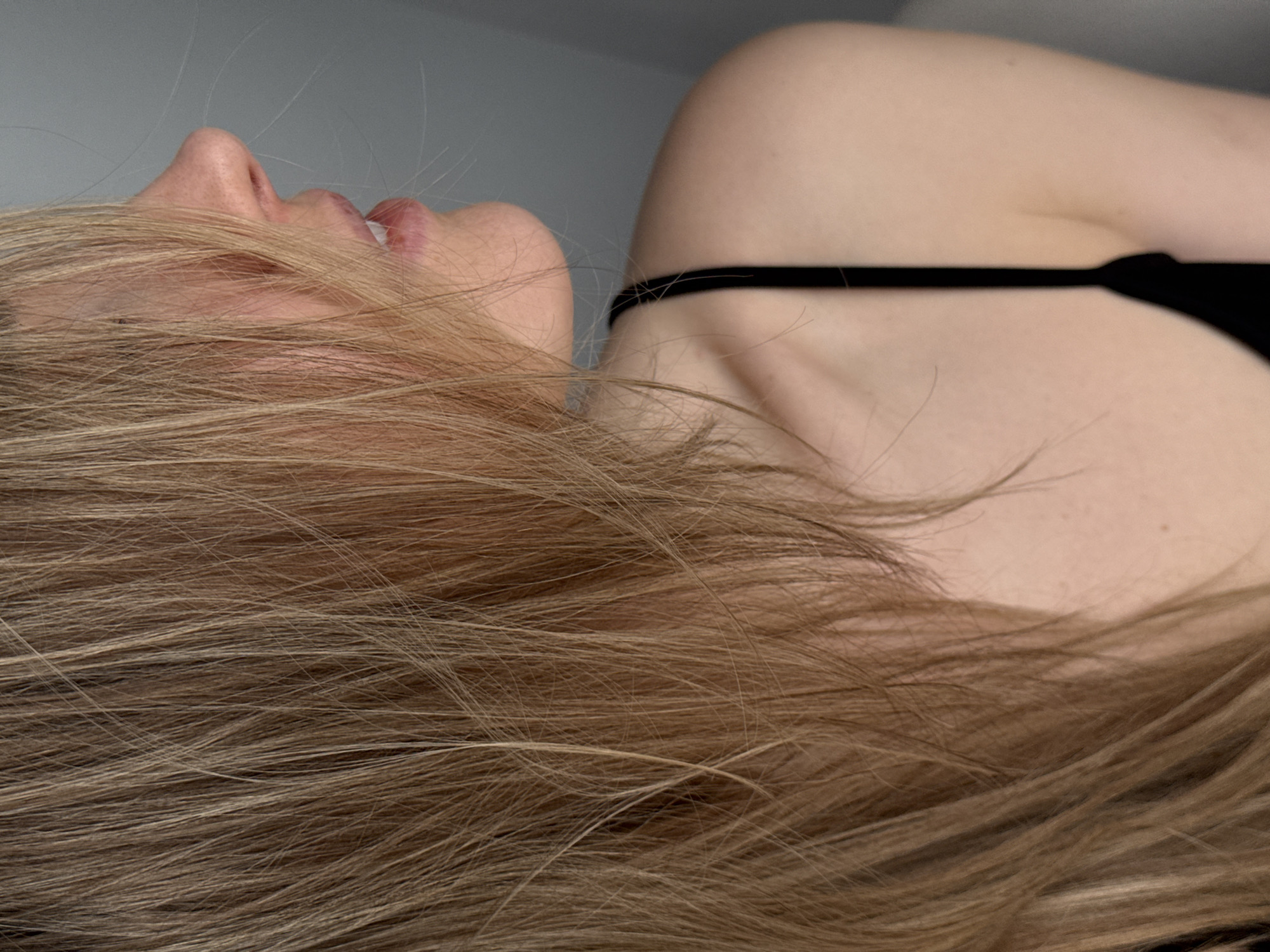 The image depicts a person with fair skin and light brown hair. The person is lying down, with their head tilted back, exposing their neck, shoulder, and side of their face. They are wearing a black garment with thin straps. The background is a plain light gray. No location in Chaikovsky, Russia, is evident or discernible in the image.