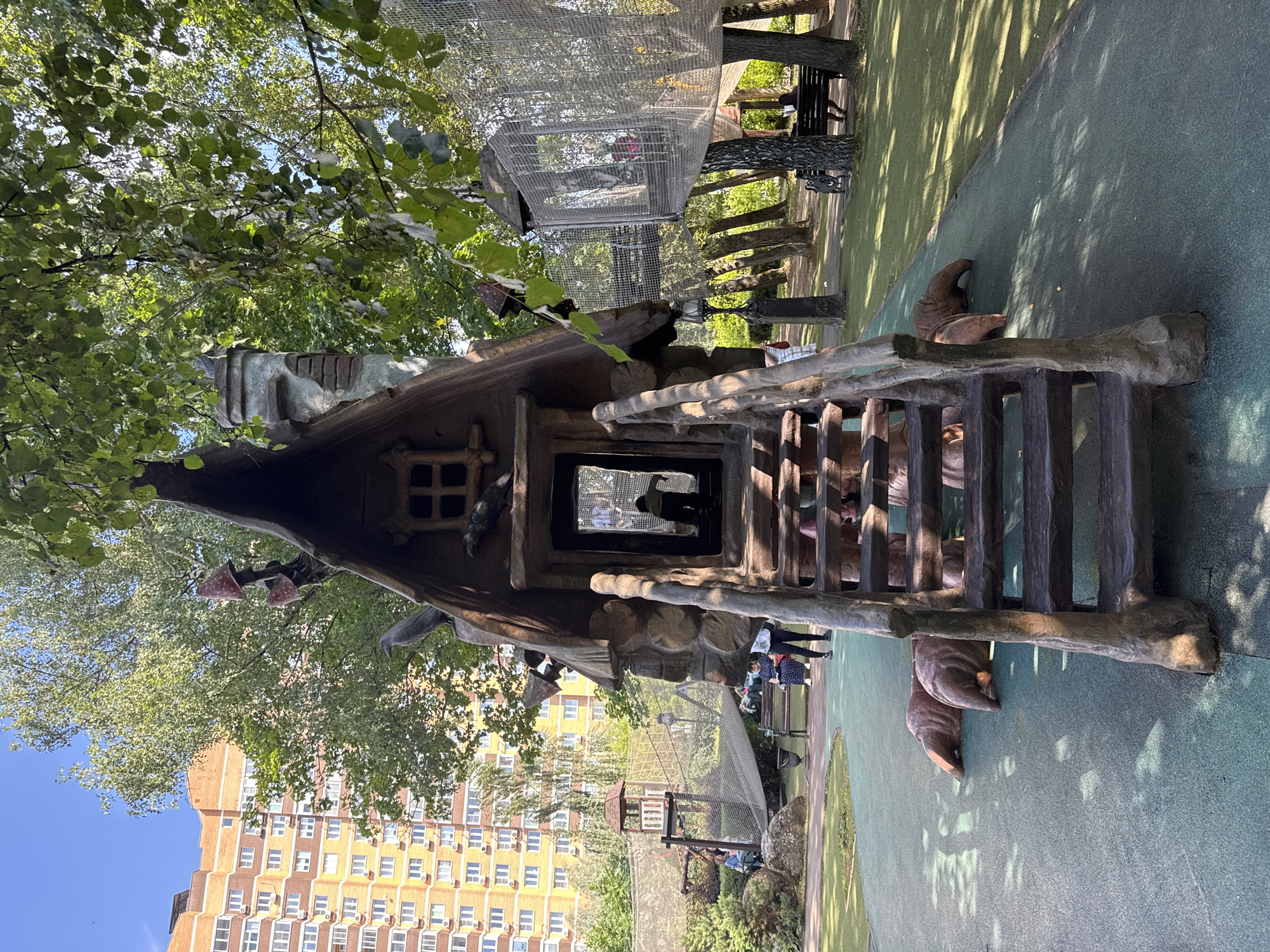 An outdoor playground with a fairytale-themed structure resembling a whimsical house is depicted. The structure features a steep, pointed roof adorned with decorative mushrooms and a faux stone chimney. A wooden staircase leads up to the entrance, which has a window and an open doorway reflecting people inside. The base of the structure is decorated with large, brown, root-like elements, possibly representing the feet or base of a mythical creature.
In the background, a tall, multi-story brick apartment building with many windows and air conditioning units is visible, characteristic of urban residential areas. Surrounding the playground is green turf and scattered trees, with dappled sunlight casting shadows. Several people are present in the scene: some are on the playground structure, while others are seated on a bench or walking in the background. A netted rope bridge structure, part of the playground's attractions, is also visible, connecting different elevated areas. The overall setting appears to be a sunny day in a park or recreational area. The location is identified as the city of Sovkhoz imeni Lenina, Russia.