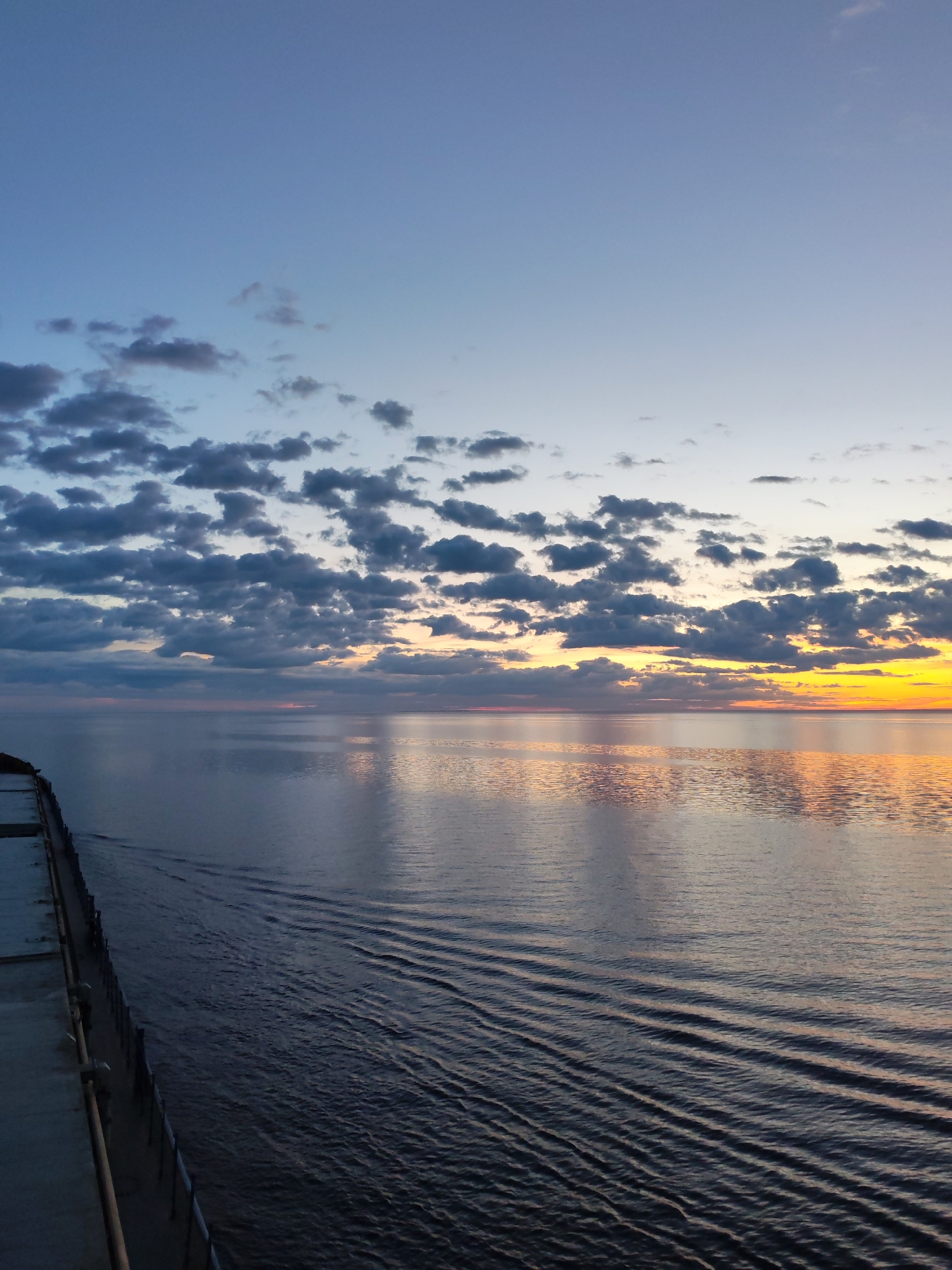 The image depicts a calm body of water under a partly cloudy sky at sunset, taken from the deck of a vessel. The sky transitions from a light blue at the top to orange and yellow hues near the horizon. The water reflects the colors of the sunset, creating a shimmering effect. Ripples radiate outwards from the vessel. The left side of the image includes part of the deck with a railing. The city of Bol'shie Mkhi, Russia is not visibly identifiable.