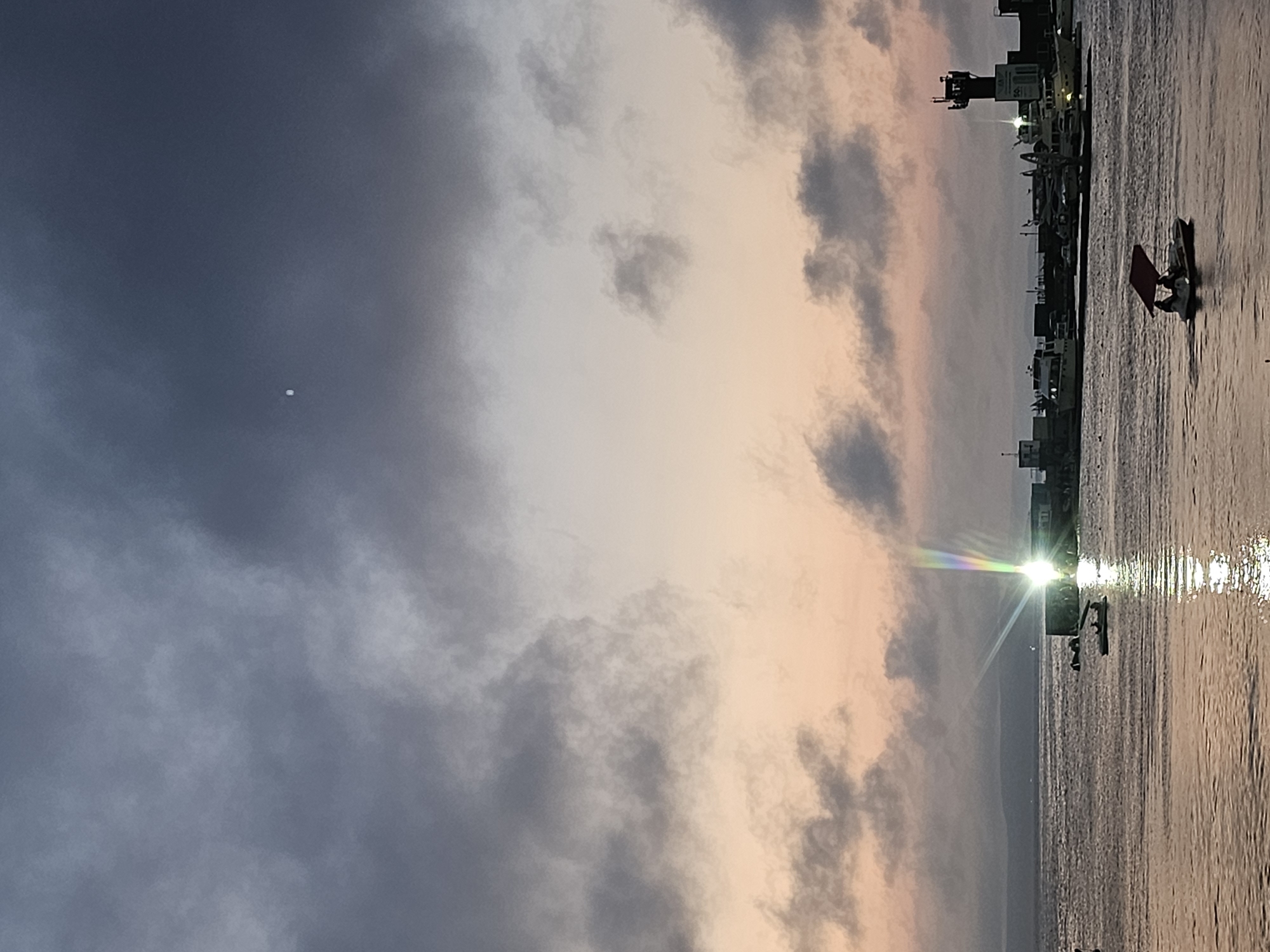 The image depicts a twilight scene at a harbor in Vladivostok, Russia. The sky is a gradient of dark gray and muted pink clouds, with a small, bright white object visible high in the sky, possibly a planet or a distant aircraft. Below the sky, a calm body of water reflects the light. In the middle ground, a marina or pier area is visible with several docked boats and a tall, dark tower with lights, likely a lighthouse or control tower. A bright light source on the pier casts a rainbow-like glare across the water. In the foreground, a small pedal boat with two people on board is moving across the water. The overall impression is one of a serene evening by the sea.