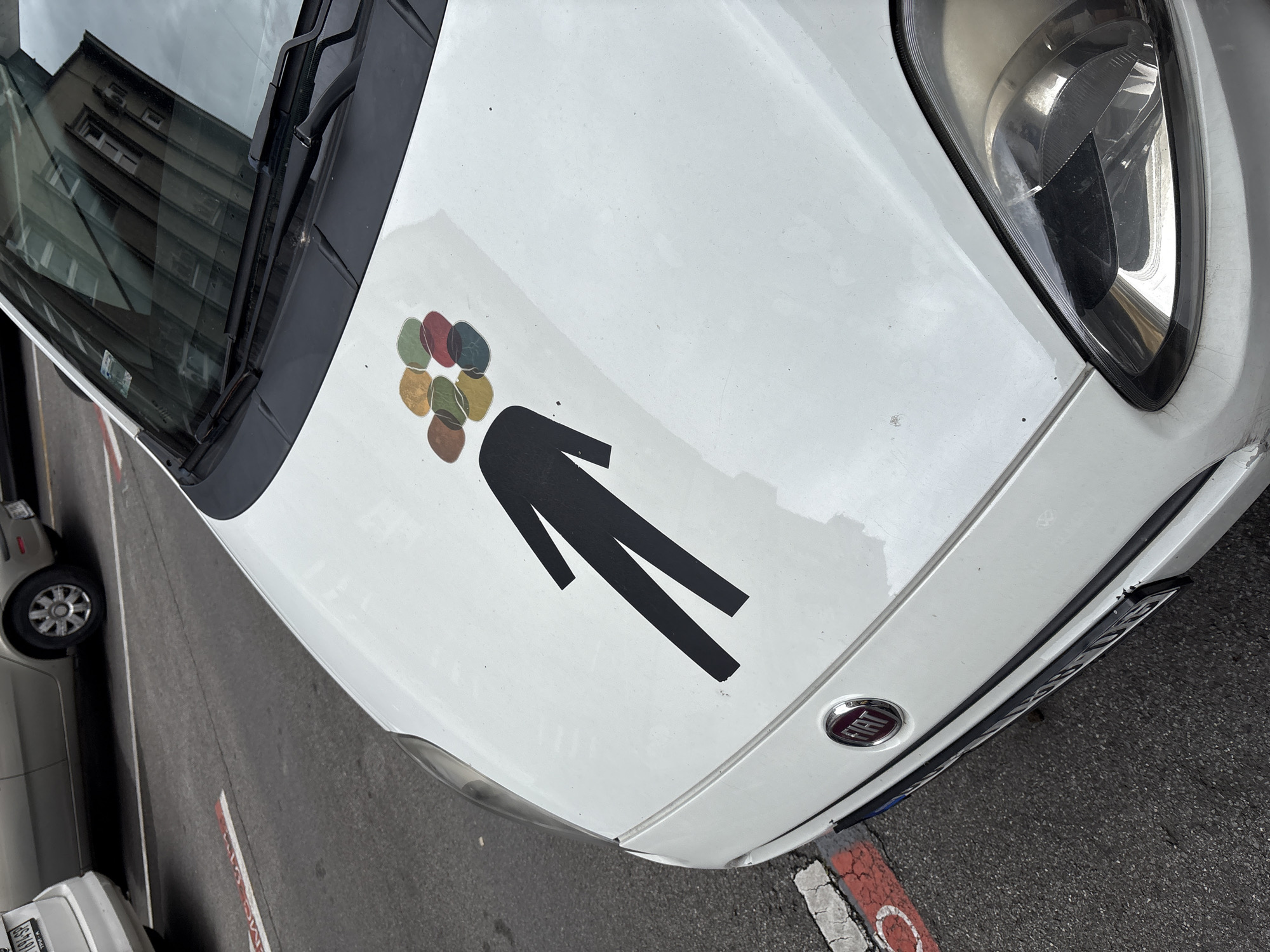 A white Fiat car is parked on an asphalt street in Belgrade, Serbia. The hood of the car features a black sticker of a human silhouette with an abstract design of colorful, overlapping circular shapes on its head. The car's logo, a red and silver oval with "FIAT" written on it, is visible on the front. Red and white painted lines are marked on the road, suggesting a parking area. The car is positioned at an angle, with its headlight and part of the windshield visible. The surrounding environment appears to be an urban setting, with other vehicles and buildings reflected in the car's windshield. The overall lighting suggests an overcast day.