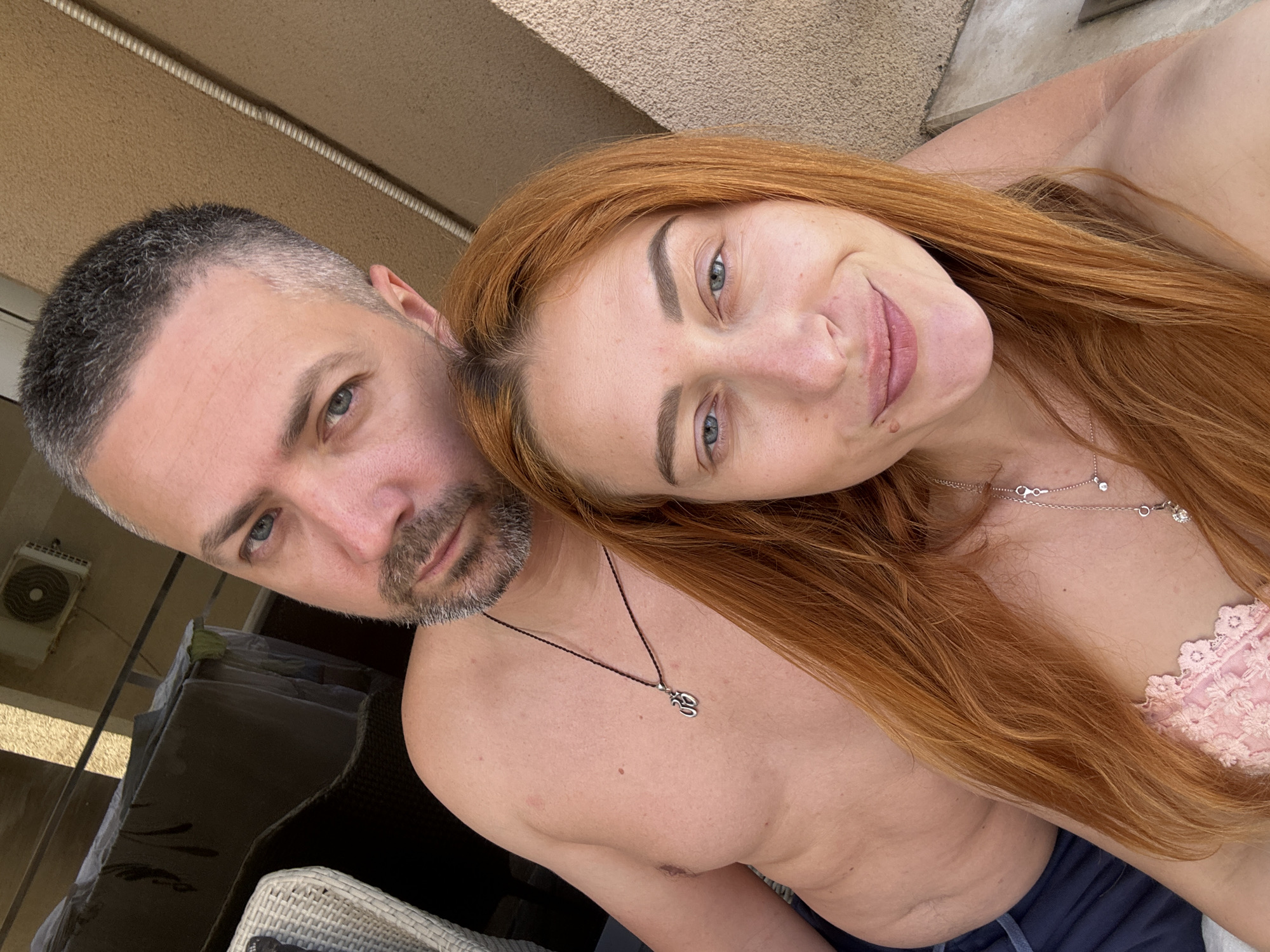A close-up selfie captures a relaxed couple outdoors, likely on a balcony or patio in Budva, Montenegro. The man, on the left, is shirtless with short, greying hair and a short beard, wearing a black cord necklace with a silver 'Om' symbol. He looks directly at the camera with intense blue-green eyes and a neutral expression.
The woman, to his right, rests her head against his shoulder. She has long, wavy reddish-brown hair and light blue eyes, with a soft smile directed at the camera. She is wearing a light pink, possibly lace or crocheted, top and a delicate silver necklace with small charms. A visible mole is on her chin.
The bright, natural lighting suggests daytime, possibly sunny. In the background, a light-colored textured wall, an air conditioning unit, and parts of outdoor wicker furniture are visible, indicating an outdoor leisure setting. There is no visible text in the image.